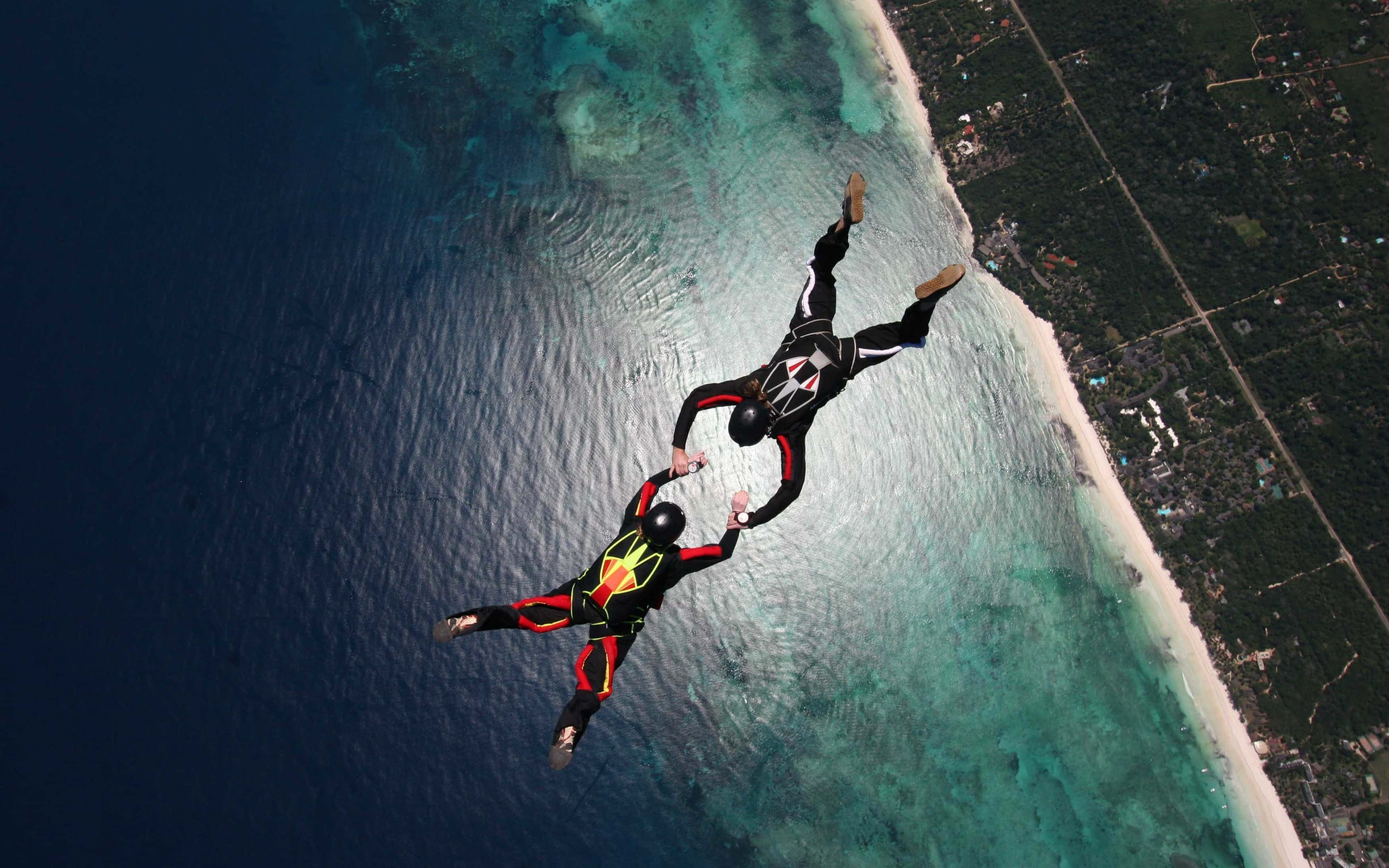 海洋 桌布 壁紙 — 兩名身穿黑色、紅色和黃色套裝的跳傘運動員在碧綠海洋和白色沙灘上空手牽手。