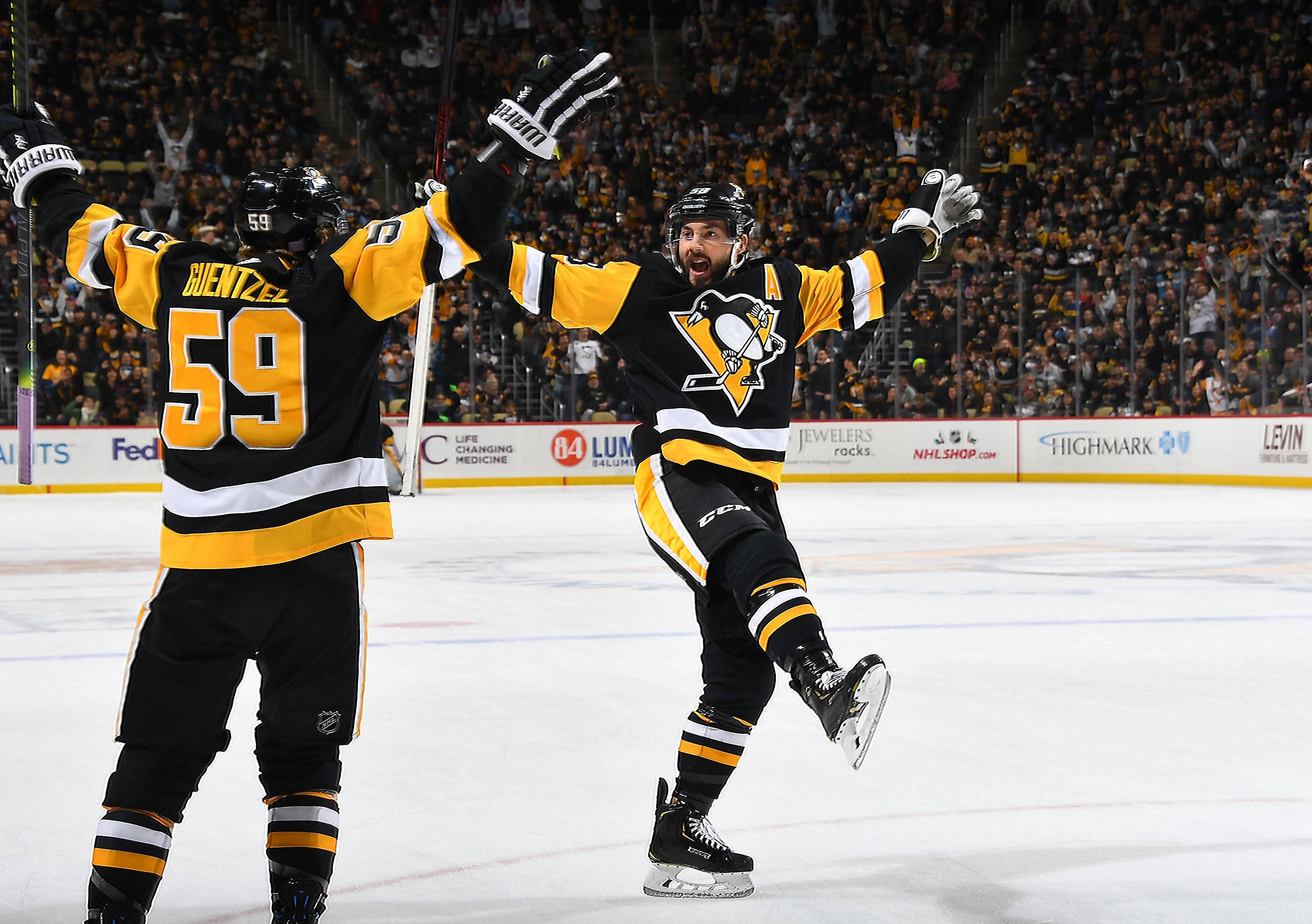 hockey 桌布 壁紙 — 兩名身穿黑色和金色球衣的曲棍球運動員在明亮的場館燈光下，在冰場上舉起手套慶祝進球。