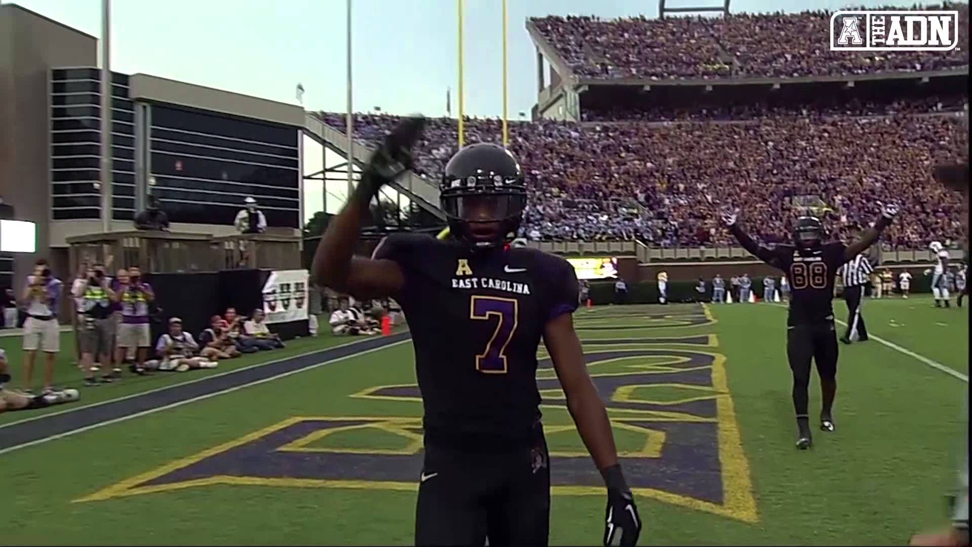 zay jones 桌布 壁紙 — 兩名身穿黑色球衣的東卡羅來納州足球員在明亮的日光下，於充滿活力的綠色球場上向歡呼的球迷揮手。