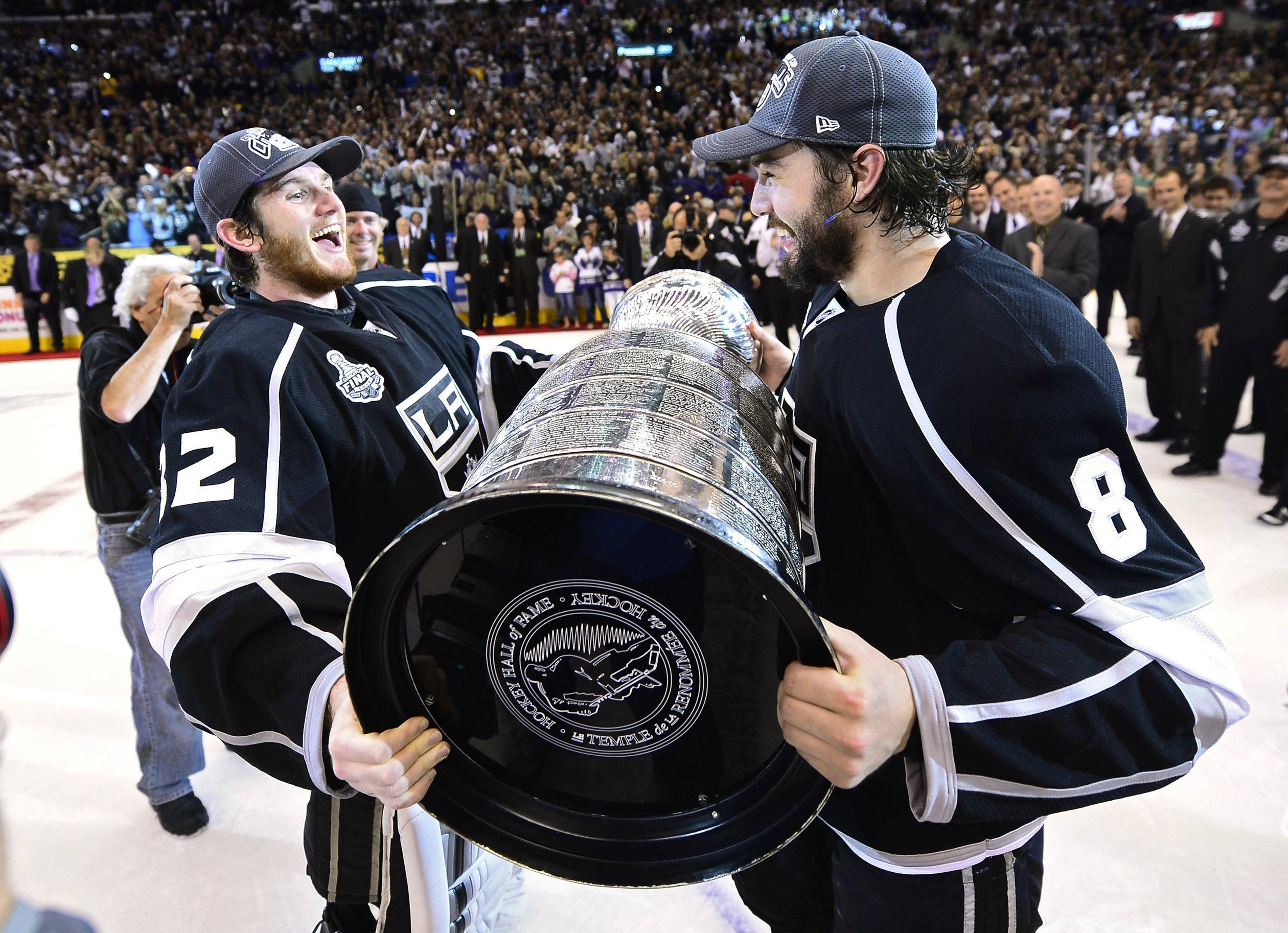 hockey 桌布 壁紙 — 兩名身穿黑色球衣的歡欣曲棍球運動員在冰場上一起高舉史丹利盃，在歡呼的人群面前。