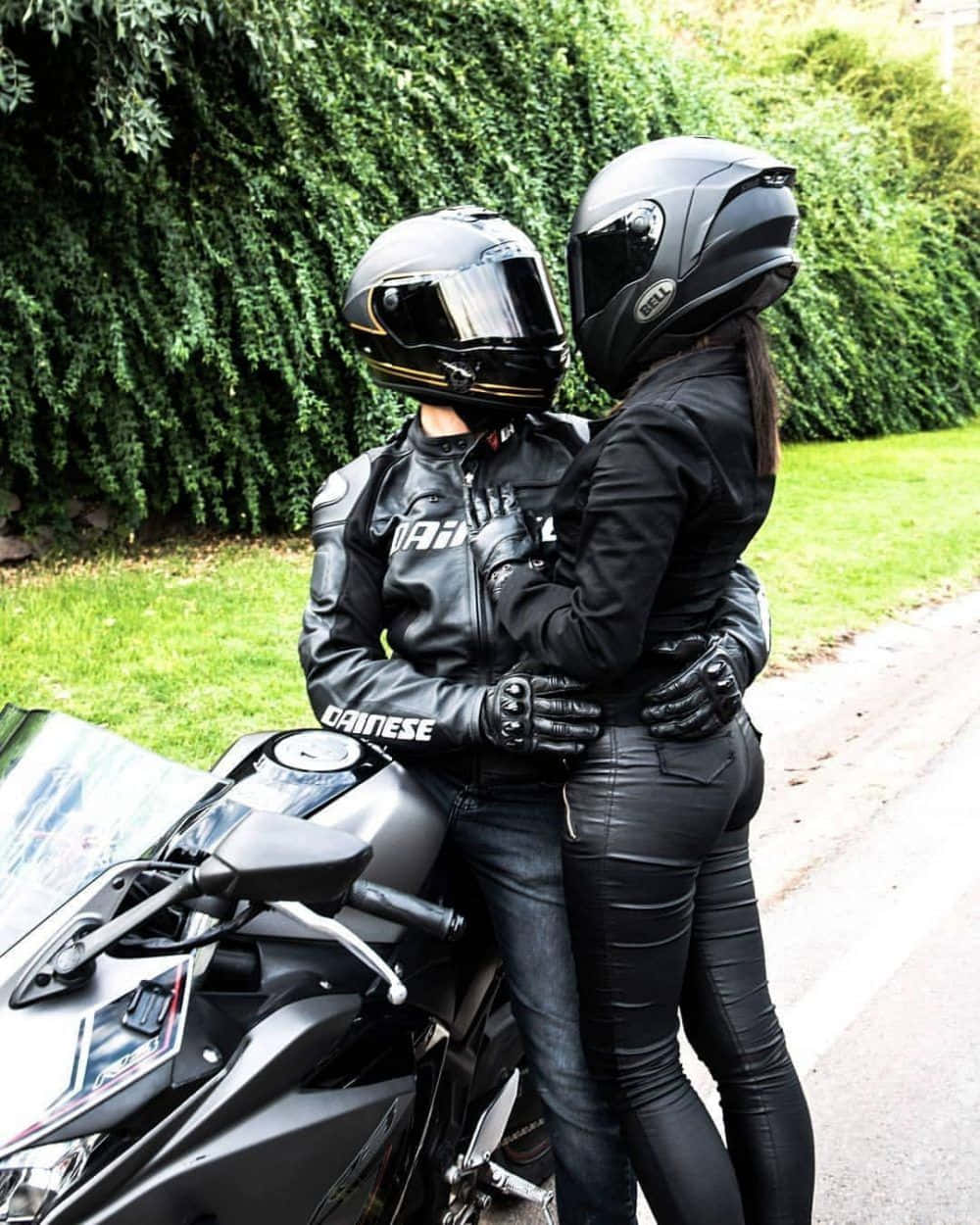 Lovers 桌布 壁紙 — 兩名身穿黑色裝備的摩托車手，在茂密綠色樹籬背景下，於一輛時尚黑色機車旁擁抱。