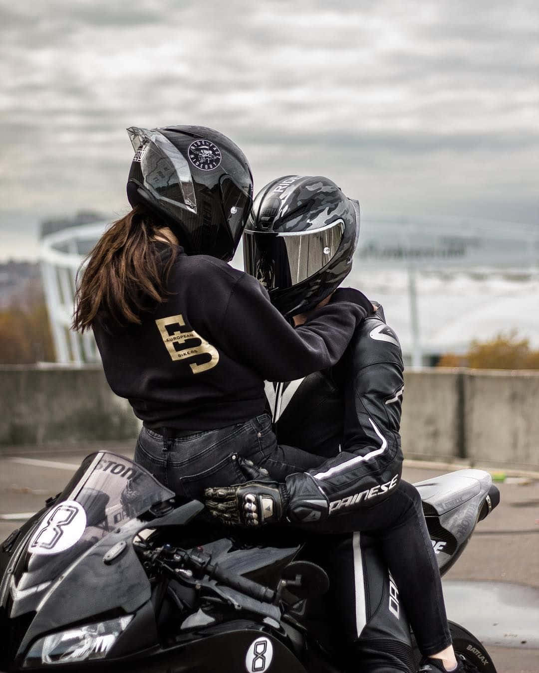 biker 桌布 壁紙 — 兩名身穿黑色裝備的摩托車手在一輛時尚的黑色摩托車上擁抱，背景是陰天和遠處的建築物。