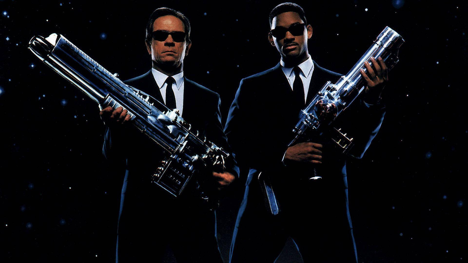 Men In Black 桌布 壁紙 — 兩名身穿黑色西裝、戴著太陽眼鏡的男子，在星空太空背景下手持未來感槍械。