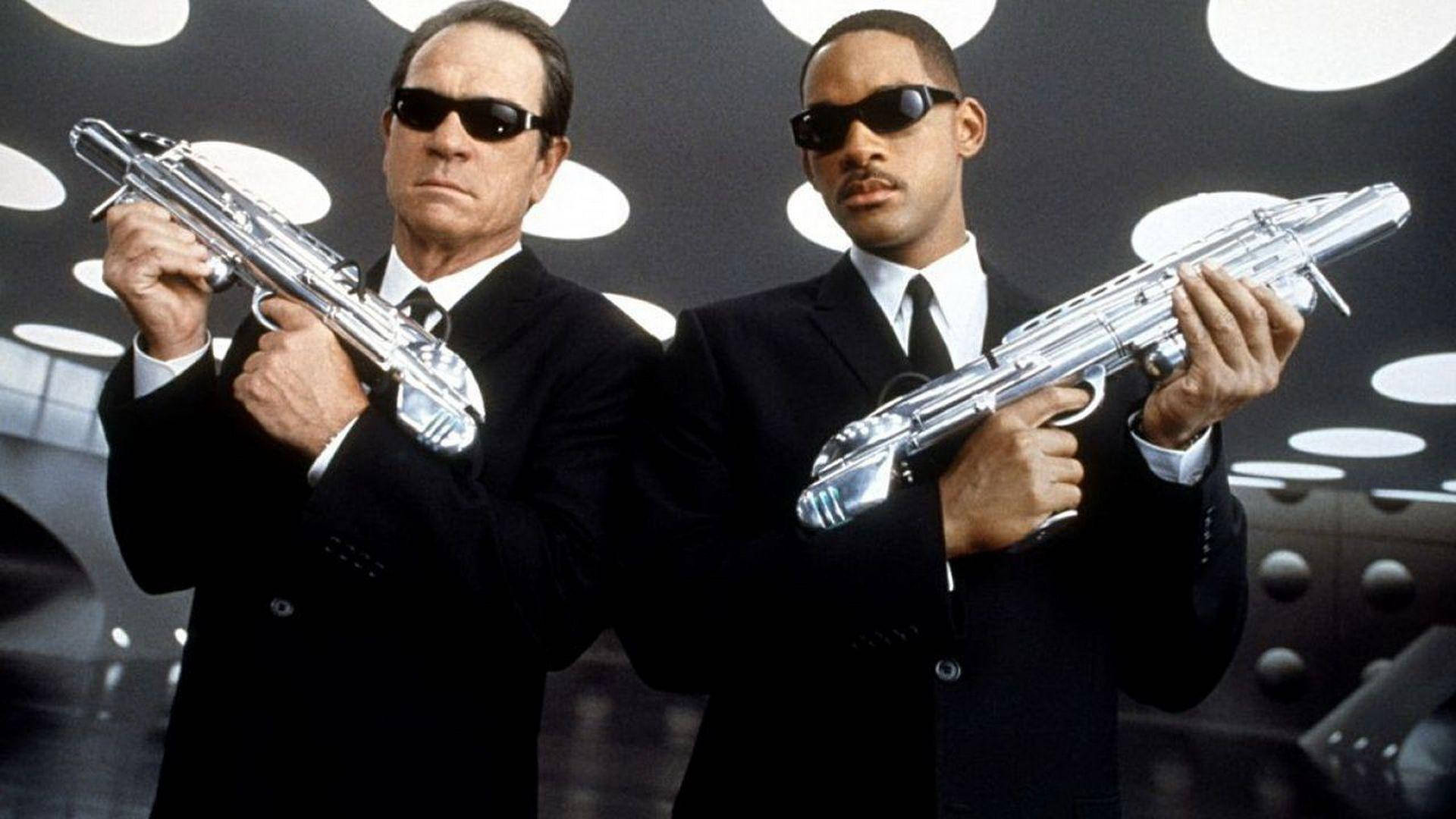 Men In Black 桌布 壁紙 — 兩名身穿黑色西裝及太陽眼鏡的男子，在發光的白色圓形燈光背景下，手持銀色未來感手槍。