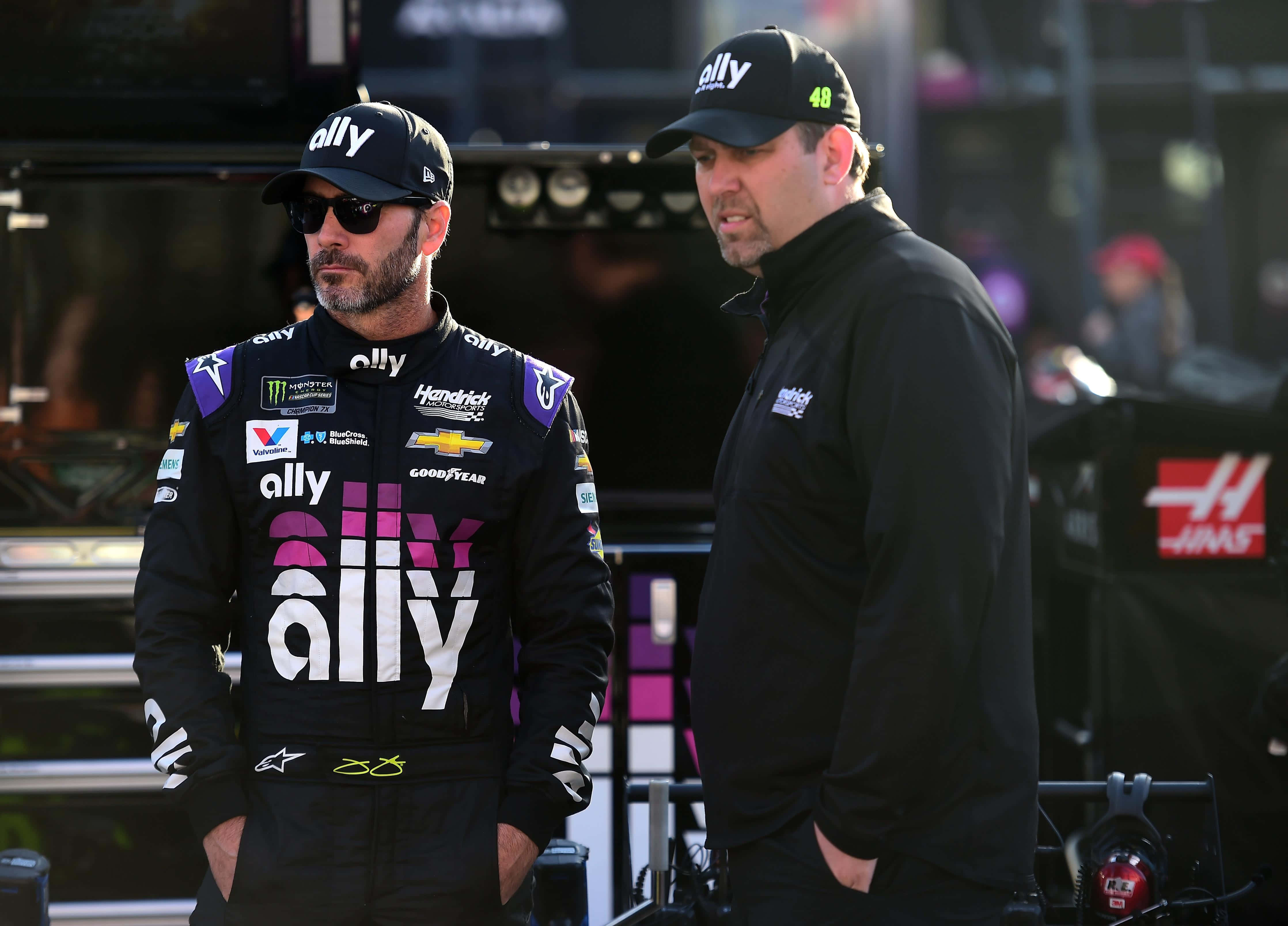 Jimmie Johnson 桌布 壁紙 — 兩名身穿黑色賽車服和帽子的NASCAR車手在白天站在維修站車庫中，贊助商標誌清晰可見。