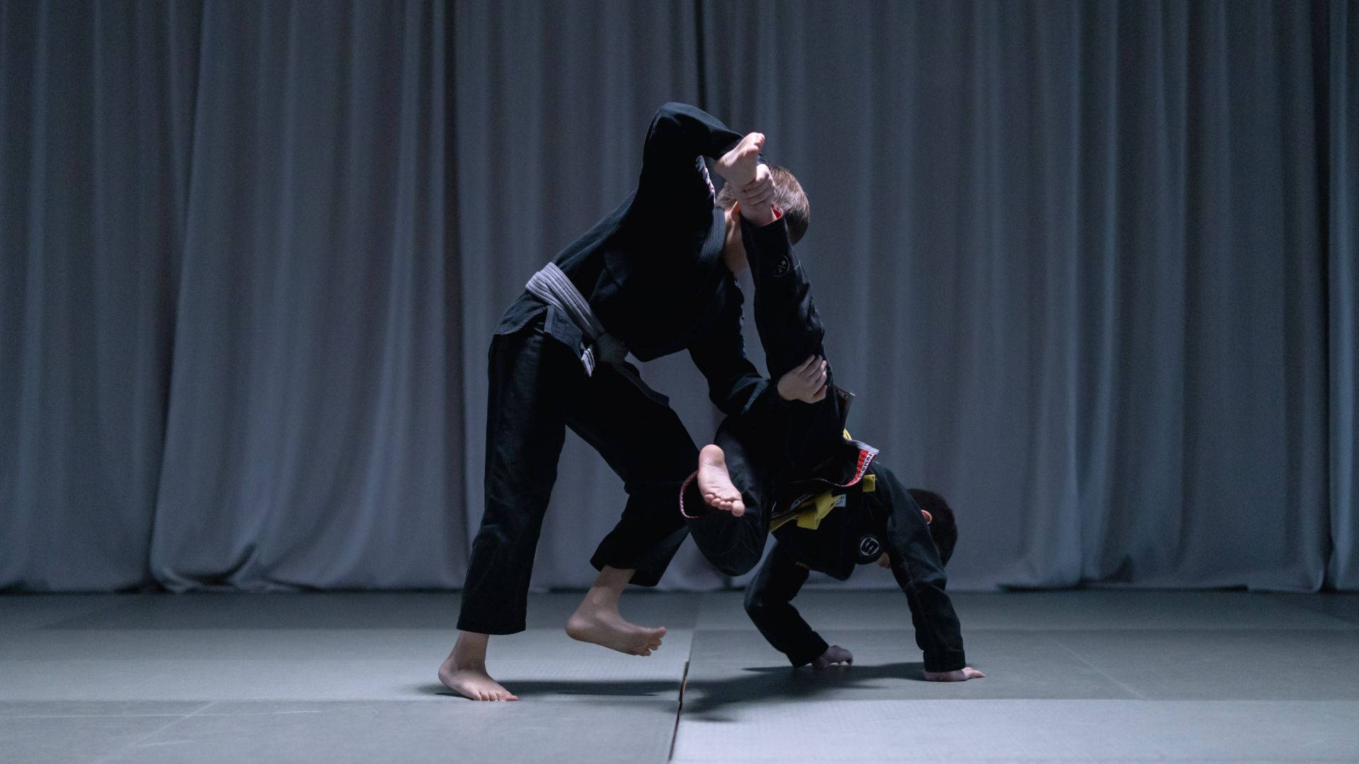 martial arts 桌布 壁紙 — 兩名身穿黑色道服的武術家在灰色軟墊上，以垂簾為背景，執行一個動態的擒拿動作。
