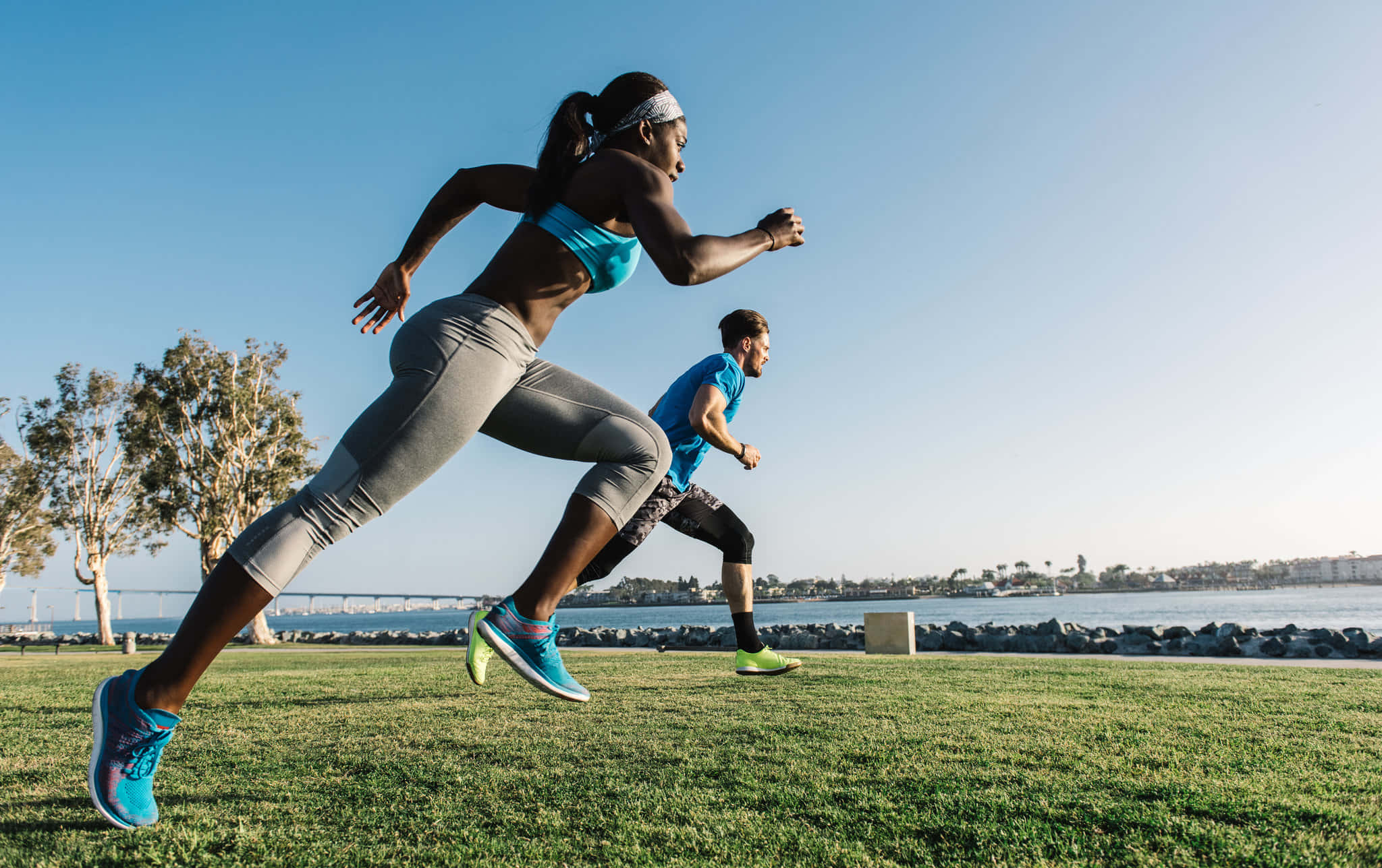Running 桌布 壁紙 — 兩名運動員在晴朗藍天下，沿著草地海濱小徑衝刺，遠處有橋樑和城市景觀。