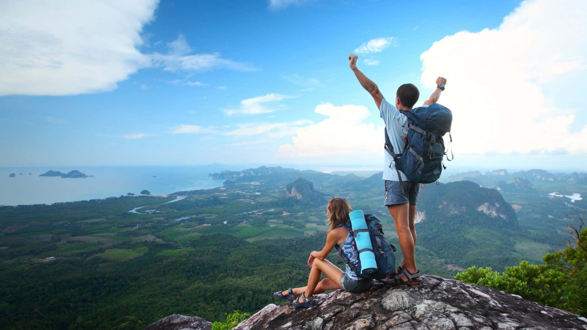 Hiking 桌布 壁紙 — 兩名遠足人士在岩石懸崖頂上慶祝，俯瞰著鬱鬱蔥蔥的綠色山谷和碧綠色的海洋，天空湛藍明亮。
