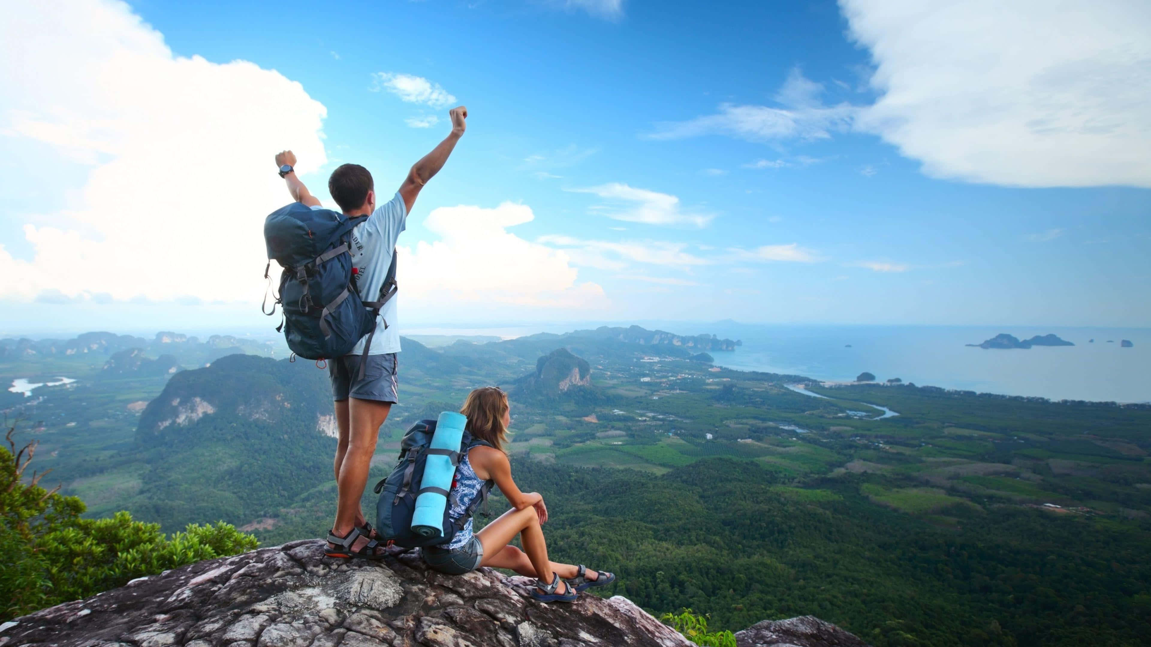 Best Travel 桌布 壁紙 — 兩名遠足者在岩石山頂上慶祝，俯瞰著鬱鬱蔥蔥的山谷和遠處的海洋，天空湛藍。