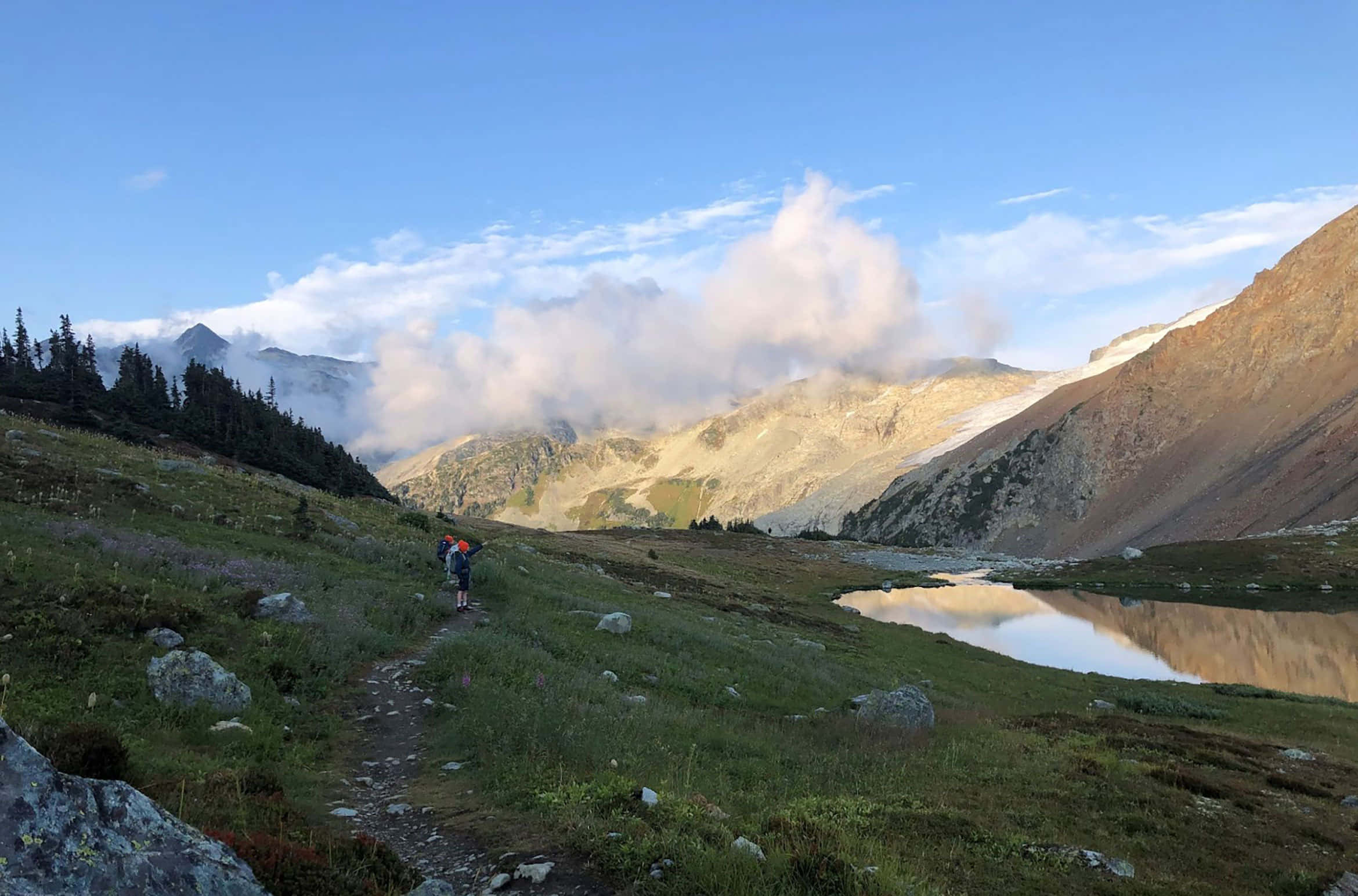 Hiking 桌布 壁紙 — 兩名遠足者沿著蜿蜒的小徑穿過陽光明媚的高山草甸，走向一個寧靜的山間湖泊，湖面倒映著金色的山峰和縷縷雲彩。