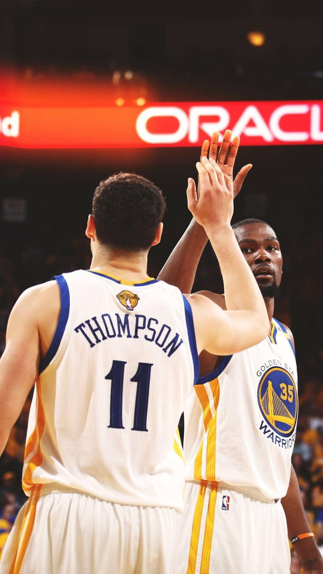 Thompson 桌布 壁紙 — 兩名金州勇士籃球運動員在燈光明亮的場館內擊掌，身後有「ORACLE」標誌發光。