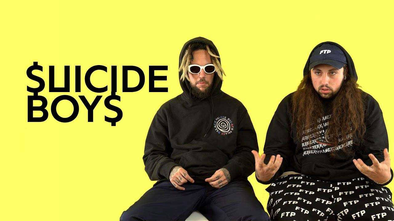 suicideboys 桌布 壁紙 — 兩名長髮戴太陽眼鏡的男子坐在明亮的黃色背景前，背景上有粗體黑色文字寫著「SUICIDE BOYS」。