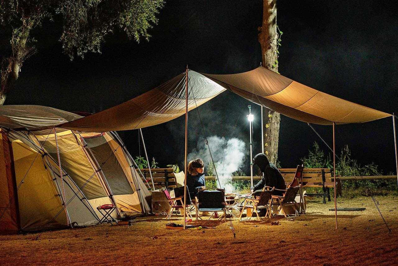Camping 桌布 壁紙 — 兩名露營者晚上坐在防水布下，在樹林地區的帳篷旁圍著冒煙的營火。