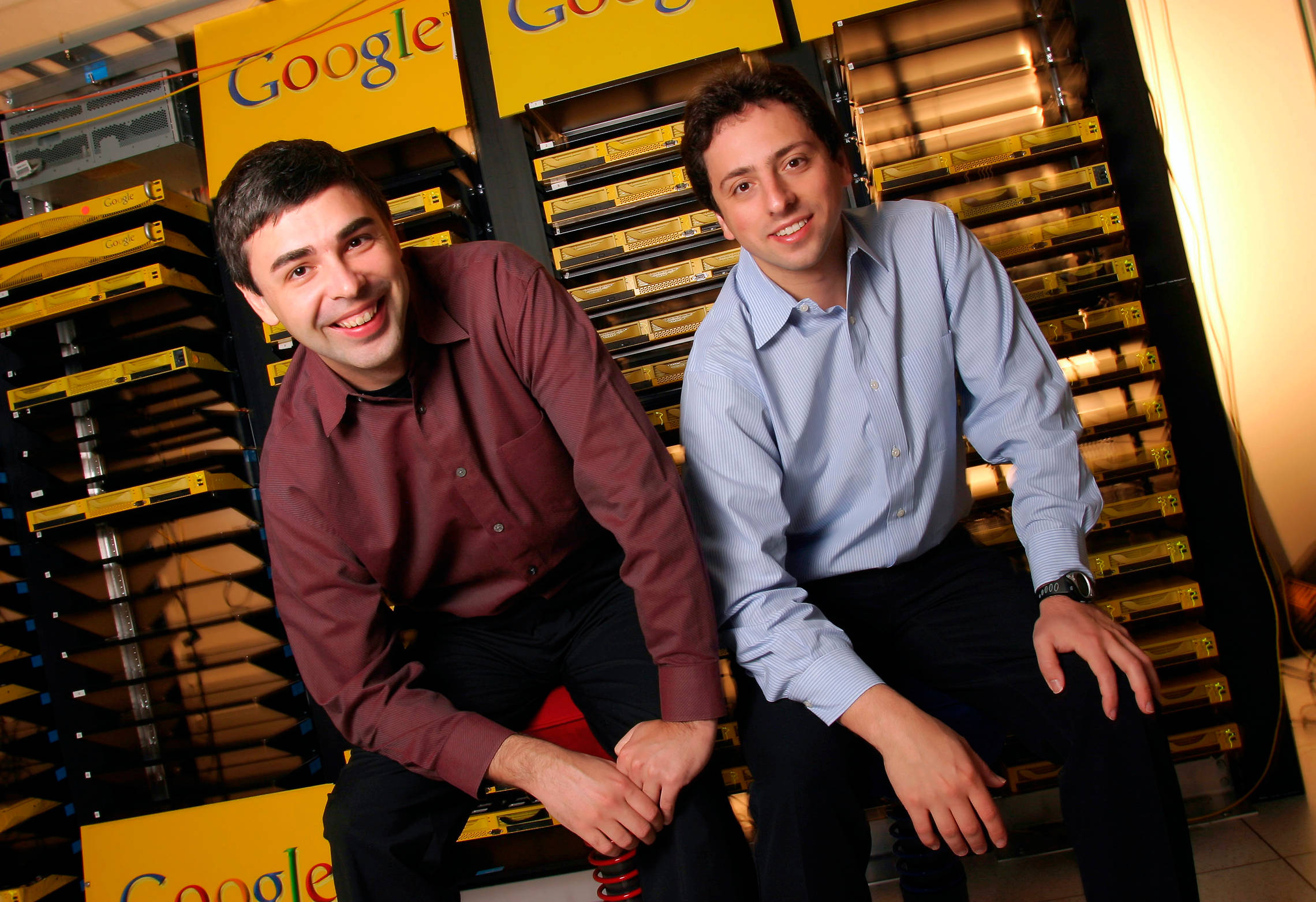 google 桌布 壁紙 — 兩名面帶微笑、身穿商務襯衫的男子，在帶有黃色品牌的Google數據中心內，隨意地坐在伺服器機架上。