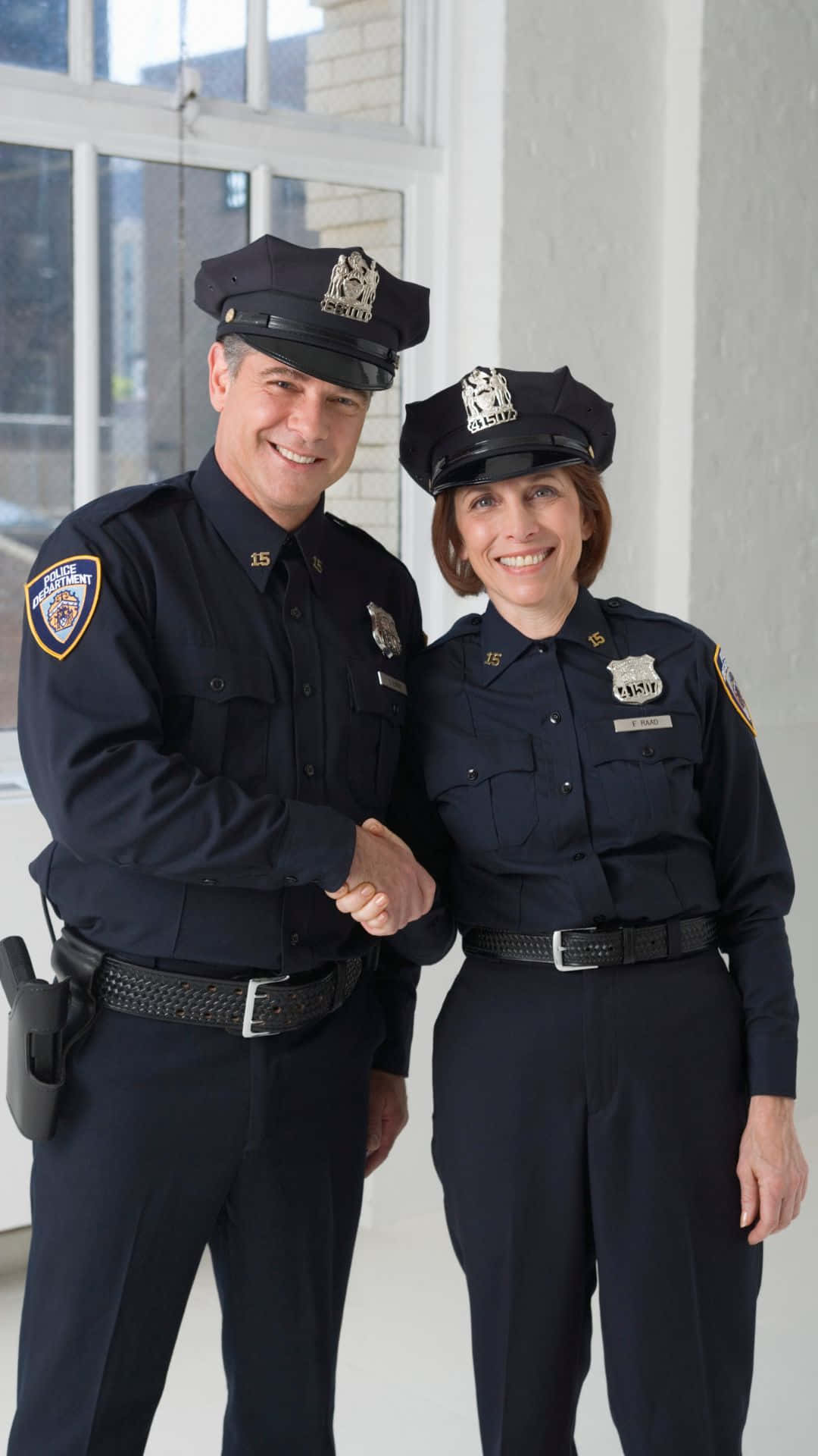 警察 桌布 壁紙 — 兩名面帶微笑、身穿深色制服的警察在室內靠近窗戶握手。