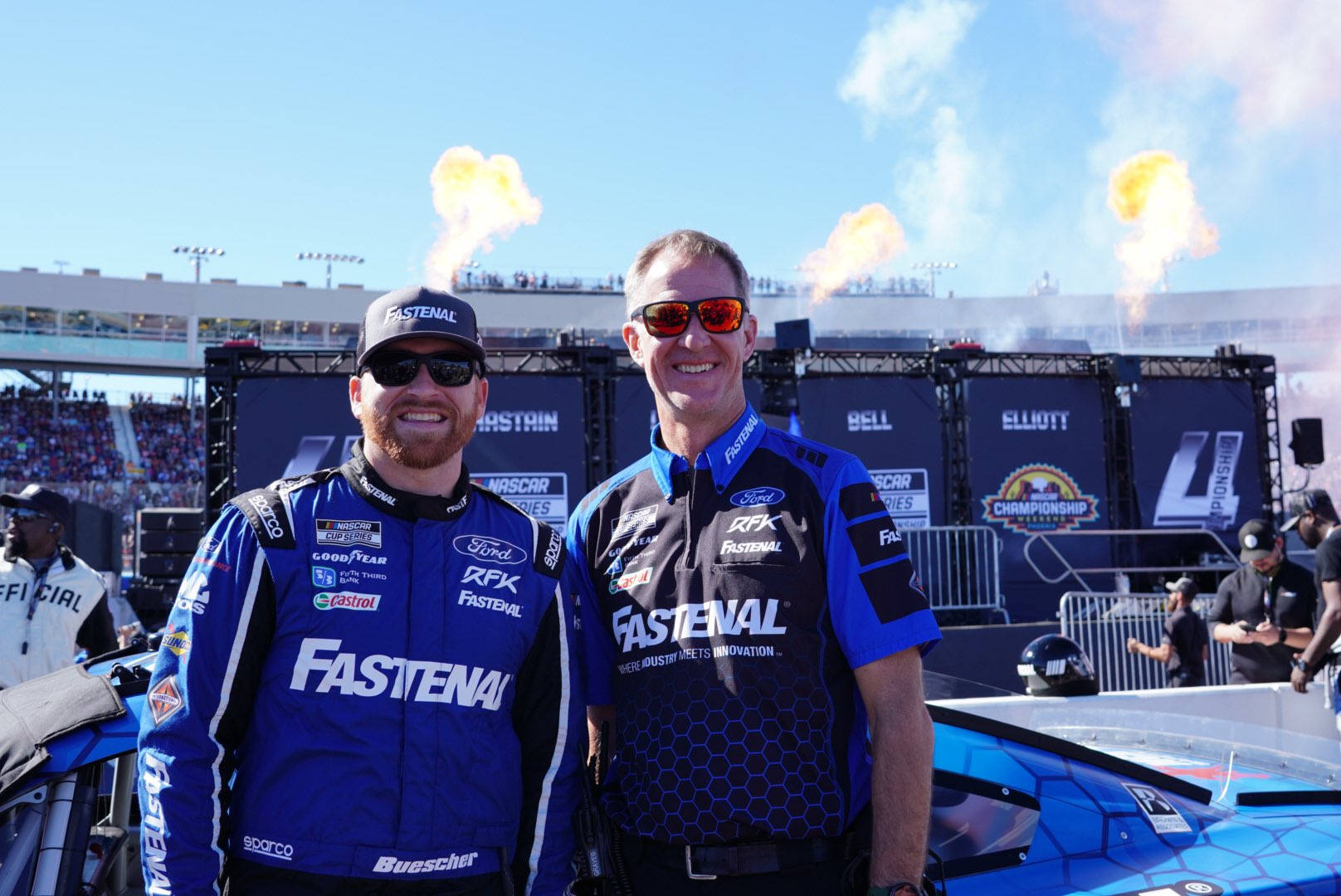 smiling 桌布 壁紙 — 兩名面帶微笑的NASCAR車手身穿藍色Fastenal賽車服，在賽車旁擺姿勢，身後有煙火爆炸。