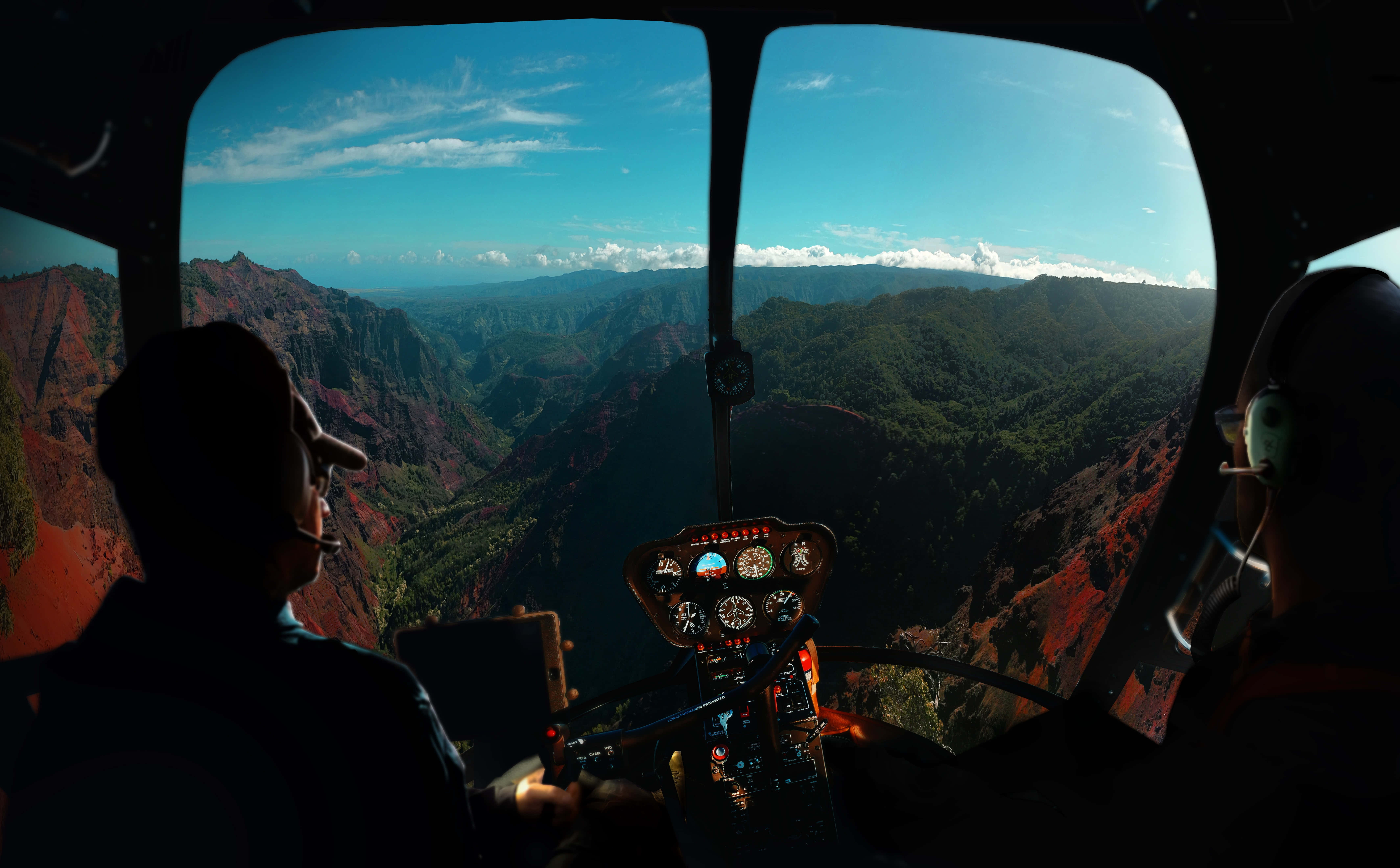 Helicopter 桌布 壁紙 — 兩名飛行員駕駛直升機飛越一個壯麗的峽谷，峽谷有紅色懸崖和綠色山谷，上方是明亮的藍天。