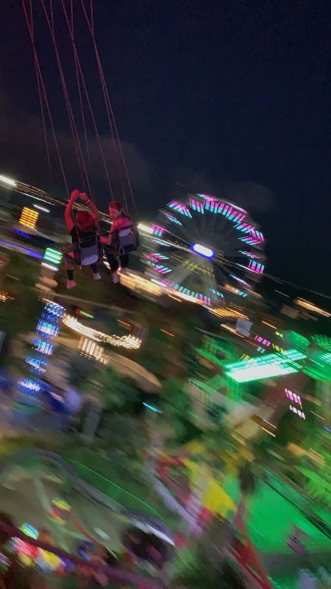 Blurry 桌布 壁紙 — 兩名騎手在充滿活力的夜間嘉年華中，在發光的摩天輪背景下，乘坐搖擺遊樂設施翱翔。