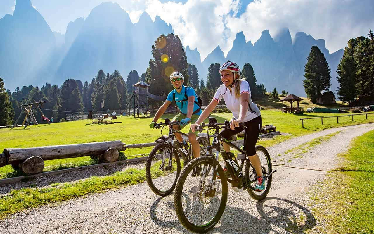Electric Bike 桌布 壁紙 — 兩名騎自行車的人在陽光明媚的碎石小徑上愉快地踩著踏板，穿過一片茂密的綠色草地，背景是高聳的鋸齒狀山脈。
