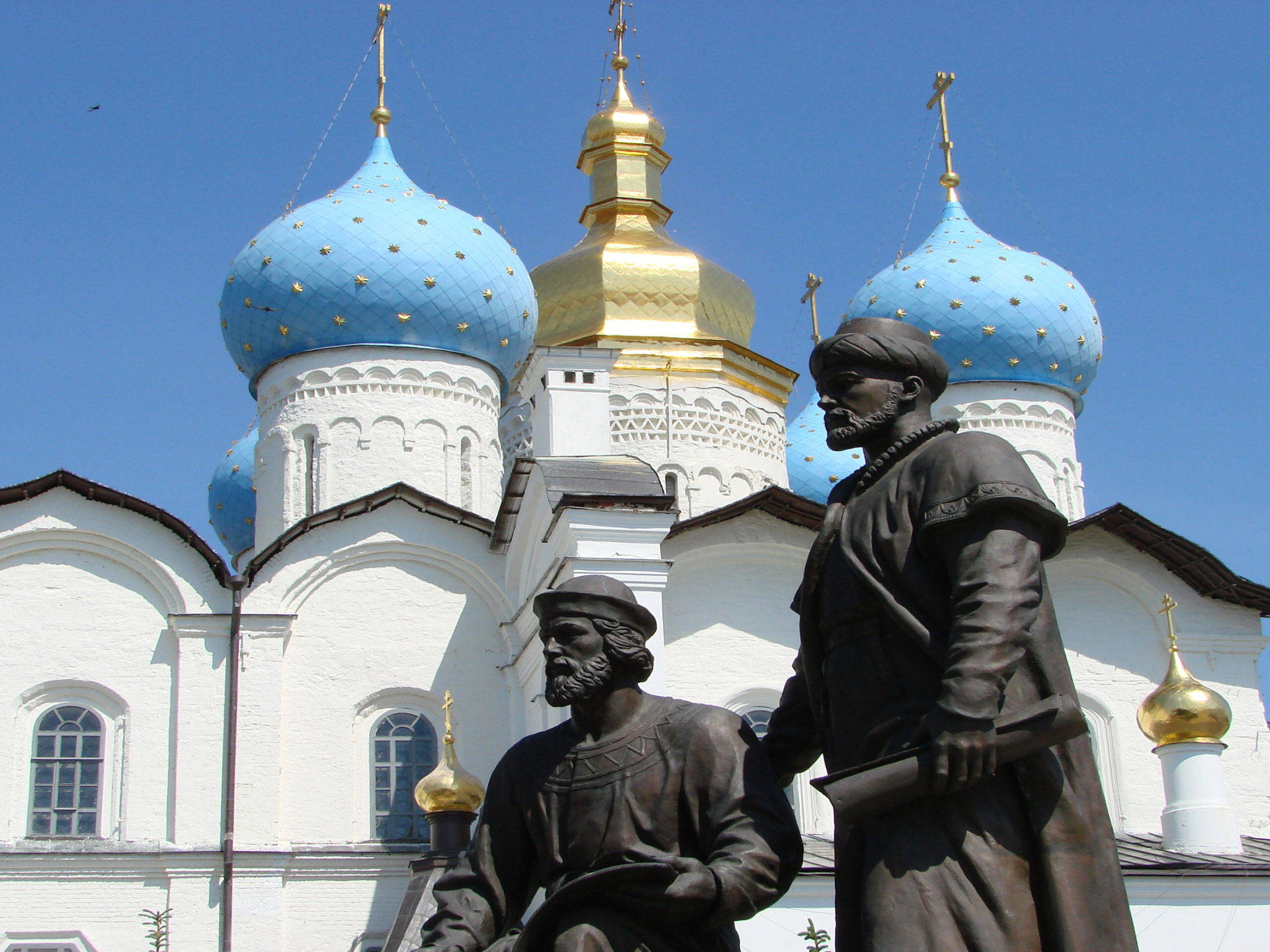 Kazan 桌布 壁紙 — 兩名鬍鬚男子的銅像矗立在一座白色教堂前，教堂有藍色和金色圓頂，背景是晴朗的藍天。