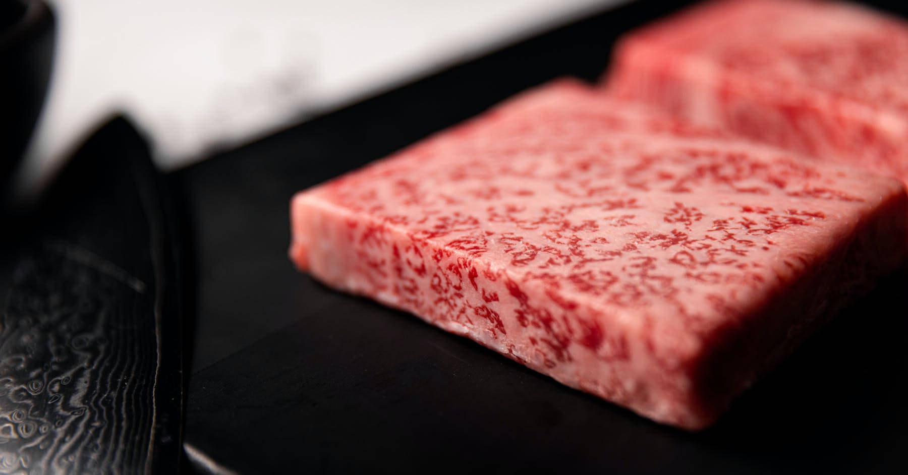 meat 桌布 壁紙 — 兩塊厚切的大理石紋和牛放在黑色盤子上，旁邊有一把帶圖案的刀。