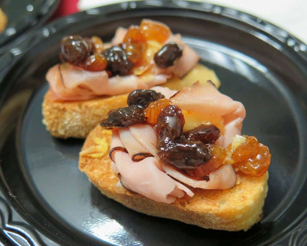 Black Forest Ham 桌布 壁紙 — 兩塊吐司麵包，上面放有火腿、葡萄乾和糖漬橙皮，放在深色盤子上。