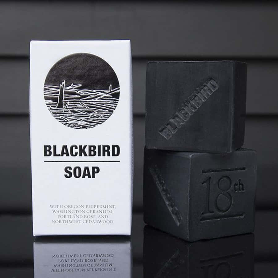Black Soap 桌布 壁紙 — 兩塊啞光黑色肥皂堆疊在一個極簡白色盒子旁邊，盒子上標有 Blackbird Soap，置於反光的深色表面上。