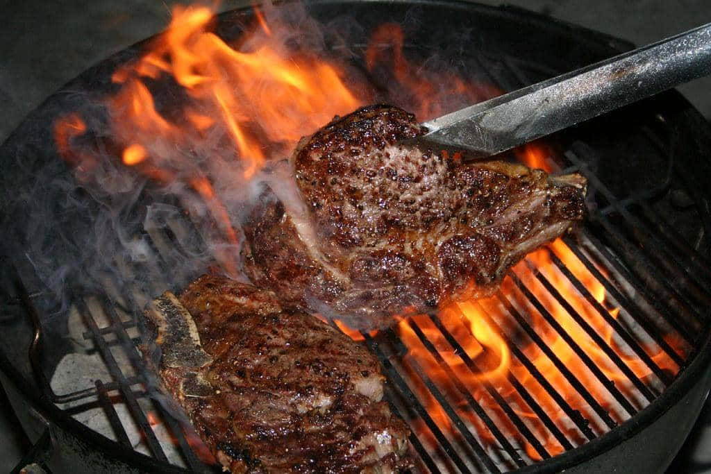 Ribeye Steak 桌布 壁紙 — 兩塊多汁牛排在黑色烤架上於熊熊烈火中滋滋作響，煙霧升騰，夾子正夾起其中一塊牛排。