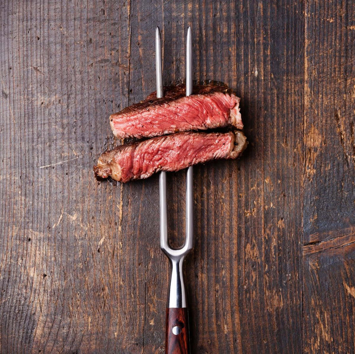 Red Meat 桌布 壁紙 — 兩塊多汁的 medium rare 牛排片用金屬叉叉住，放在深色鄉村風木質表面上。