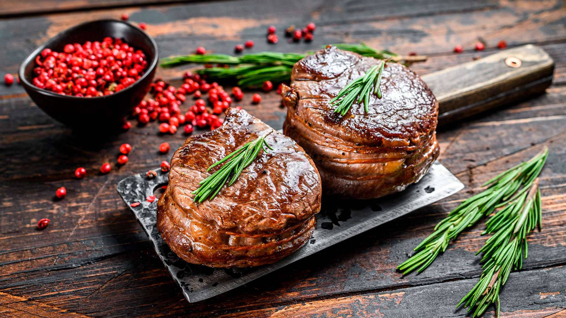 Food Photography 桌布 壁紙 — 兩塊多汁的烤牛肉排以迷迭香點綴，放在一塊石板盤上，旁邊有一碗紅胡椒粒，置於鄉村風木桌上。