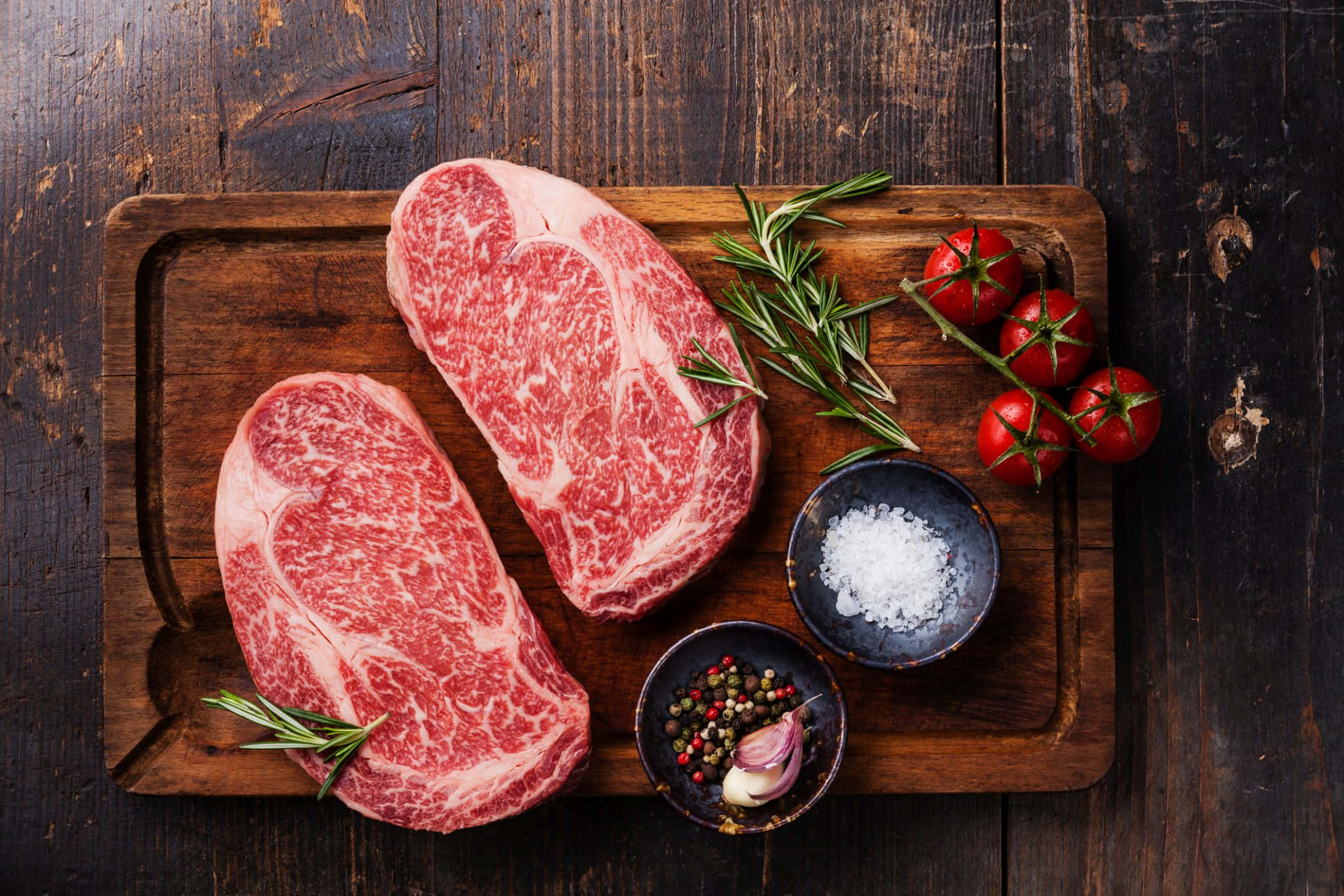 meat 桌布 壁紙 — 兩塊大理石紋牛排放在質樸的木板上，搭配迷迭香、櫻桃番茄、鹽和胡椒粒。