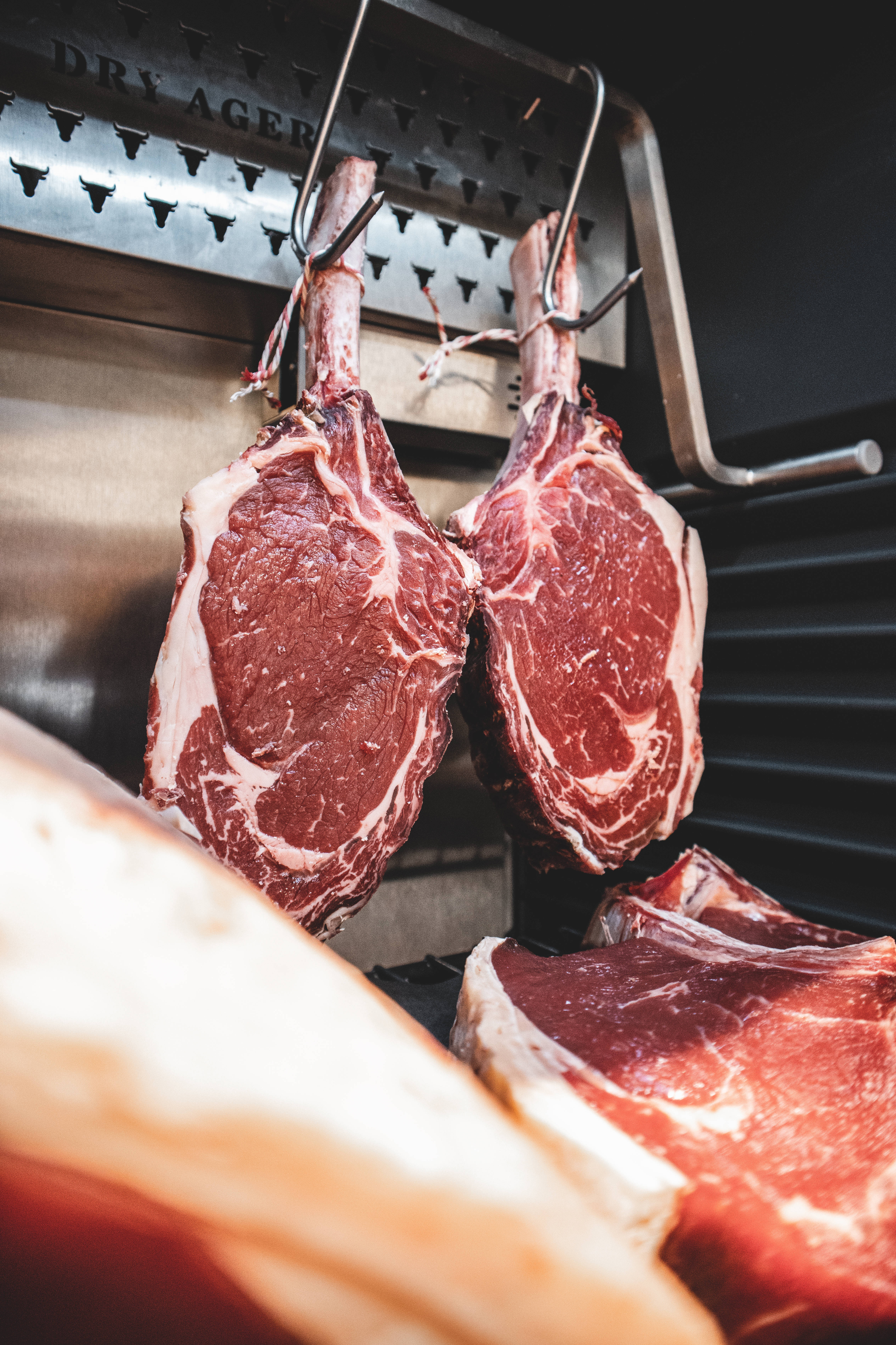 meat 桌布 壁紙 — 兩塊大理石紋牛肉斧頭扒掛在不鏽鋼乾式熟成室的掛鉤上，下方有其他切塊。