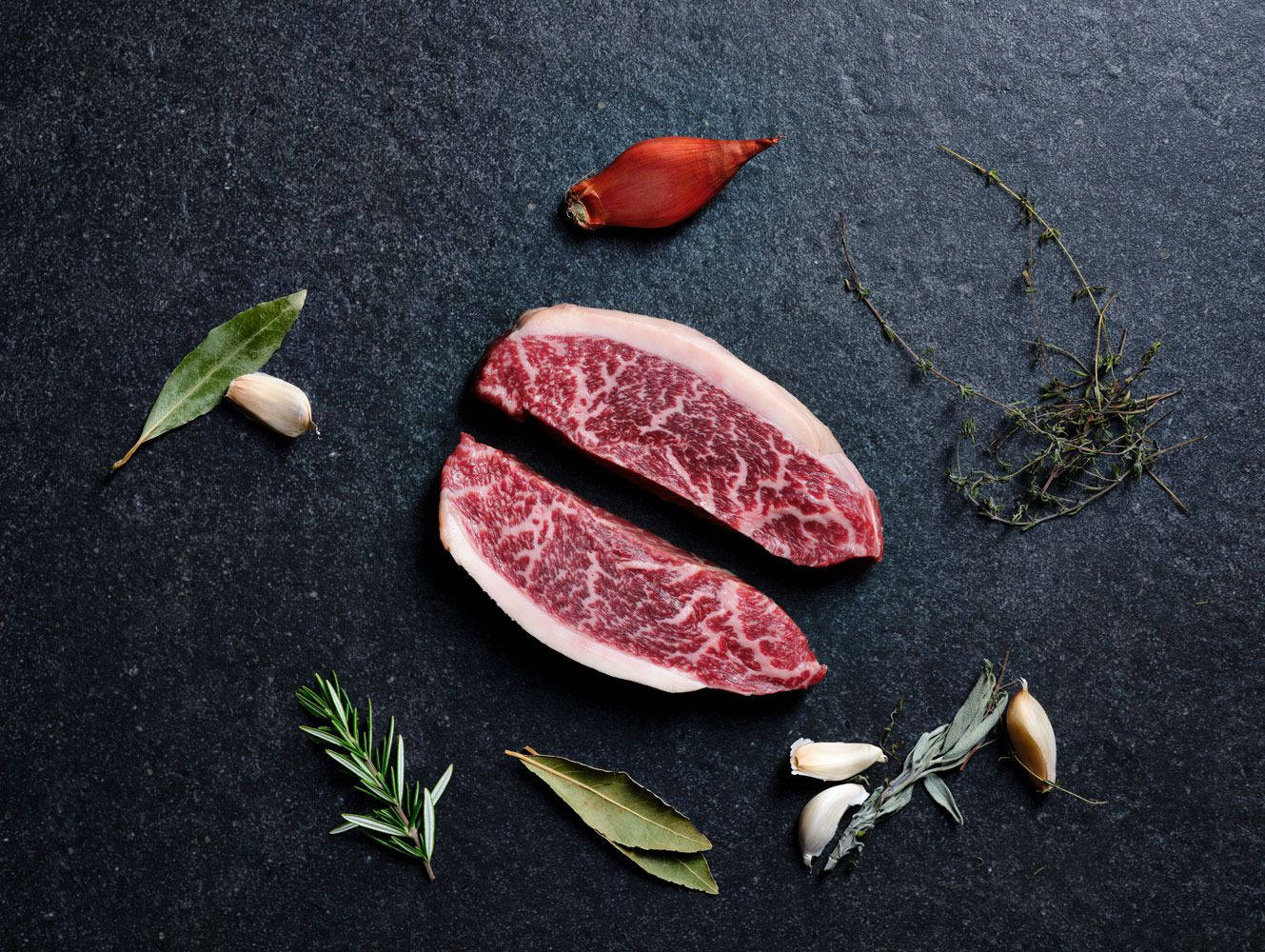 meat 桌布 壁紙 — 兩塊大理石紋牛肉牛排放在深色石板表面上，周圍是新鮮香草和蒜瓣。
