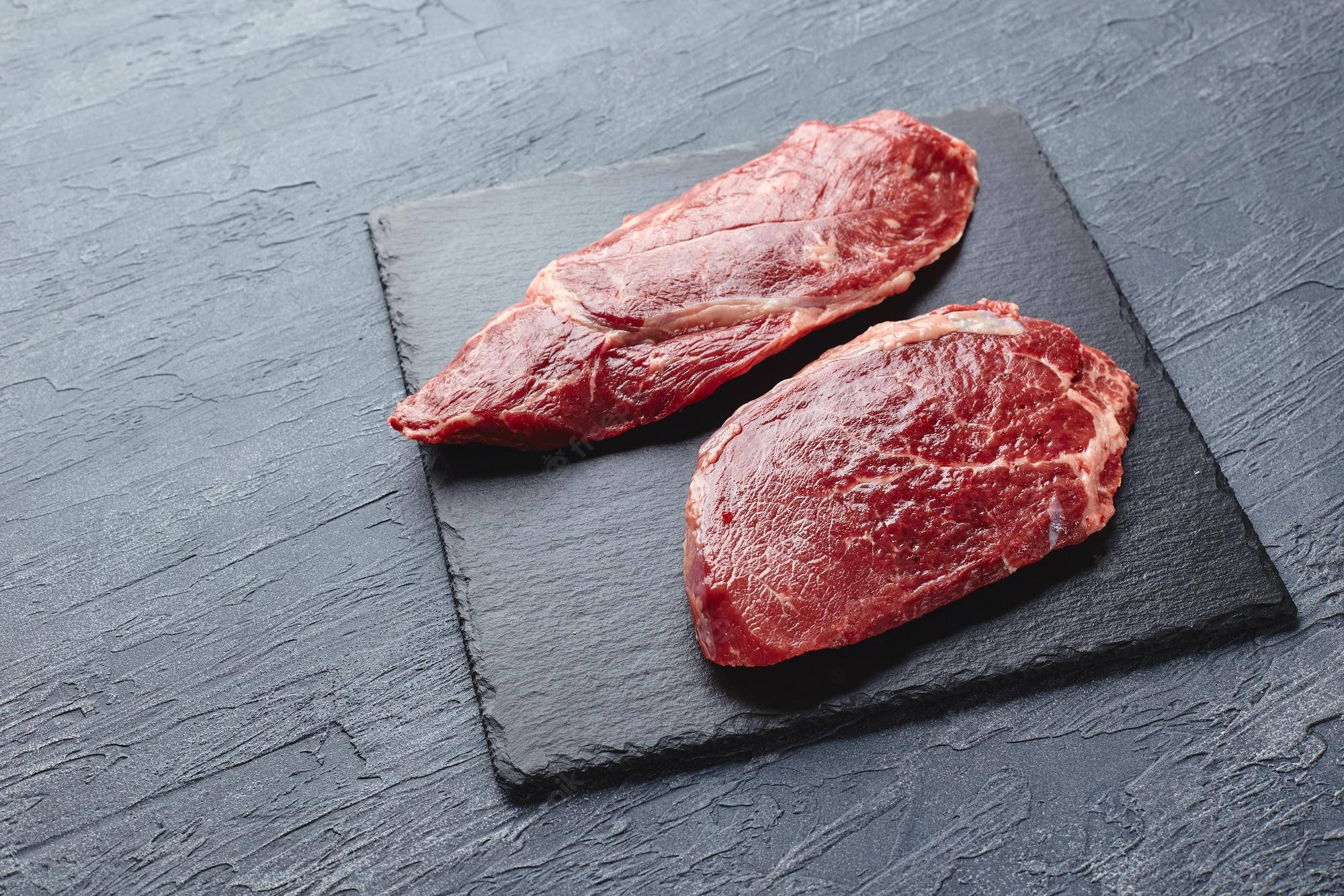 meat 桌布 壁紙 — 兩塊帶有大理石紋的未經烹調的牛排放在深色石板板上，背景為有紋理的灰色。