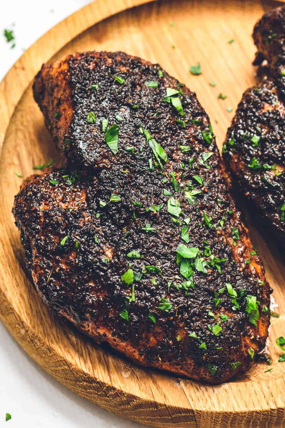 Blackened Chicken 桌布 壁紙 — 兩塊帶有深色香草外皮和新鮮巴西里的香煎雞胸肉，放在質樸的木盤上。