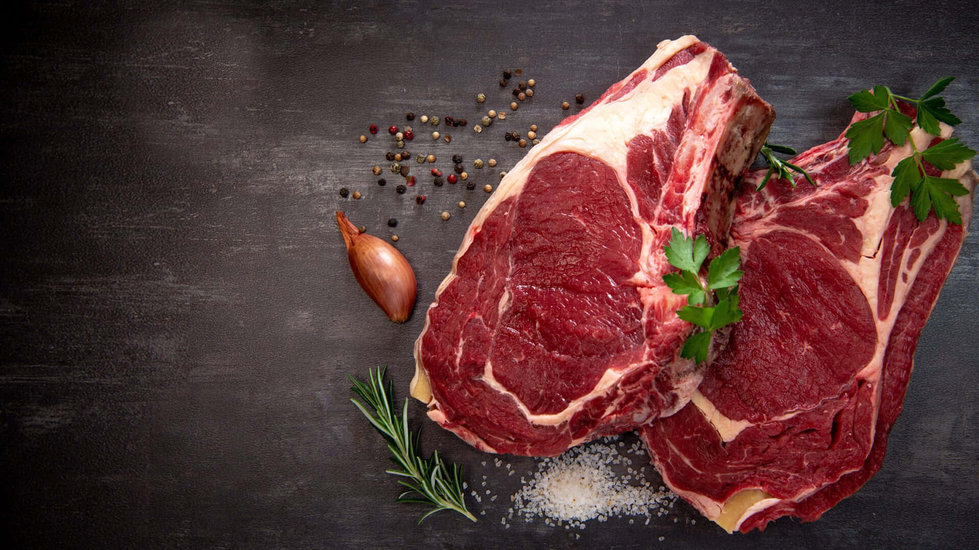Red Meat 桌布 壁紙 — 兩塊帶骨生肋眼牛排，以迷迭香、歐芹、鹽和胡椒粒裝飾，放在深色質樸的表面上。