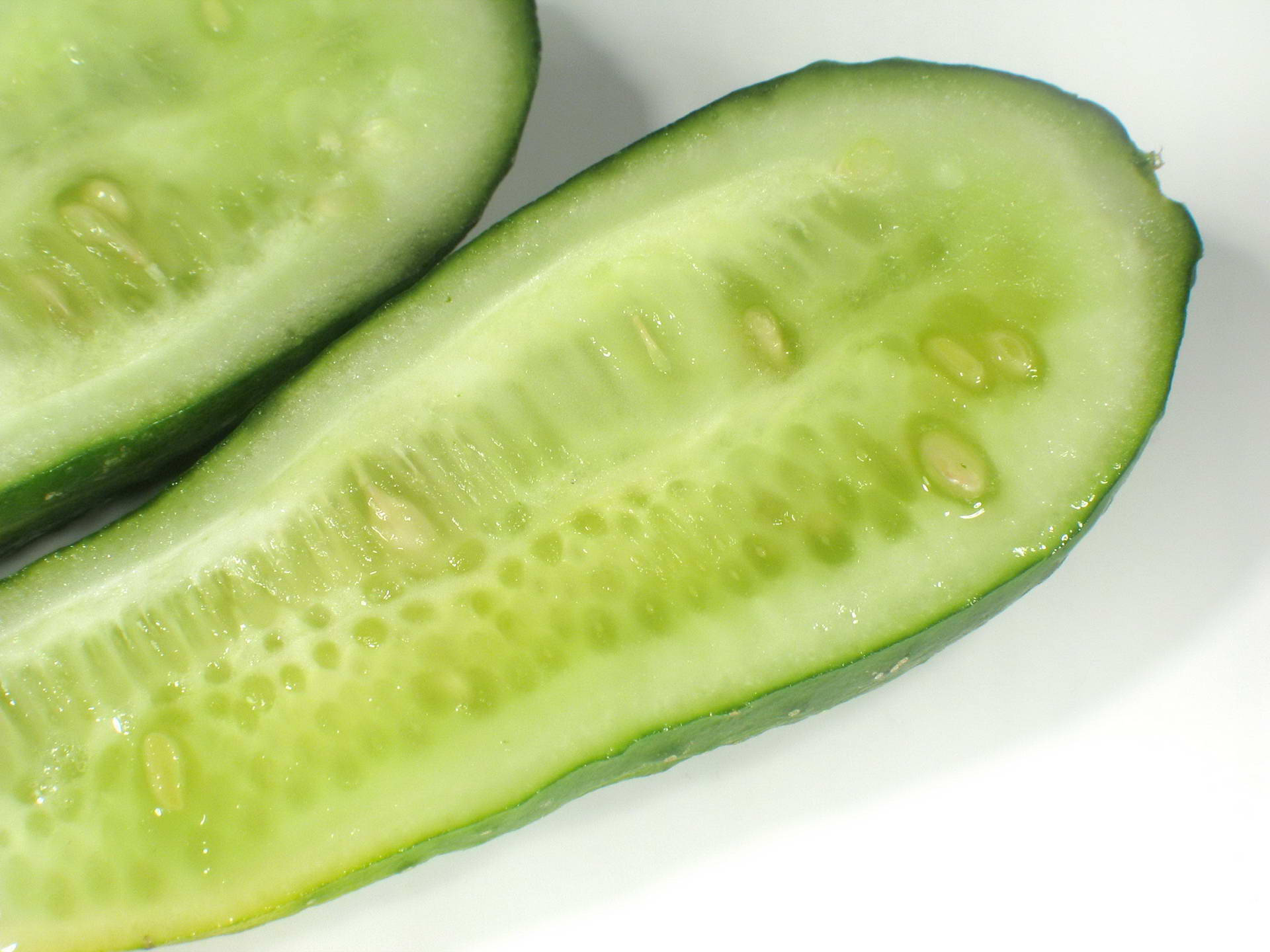 sliced 桌布 壁紙 — 兩塊新鮮小黃瓜切半的特寫，露出淡綠色果肉和細小種子，背景為純白色。