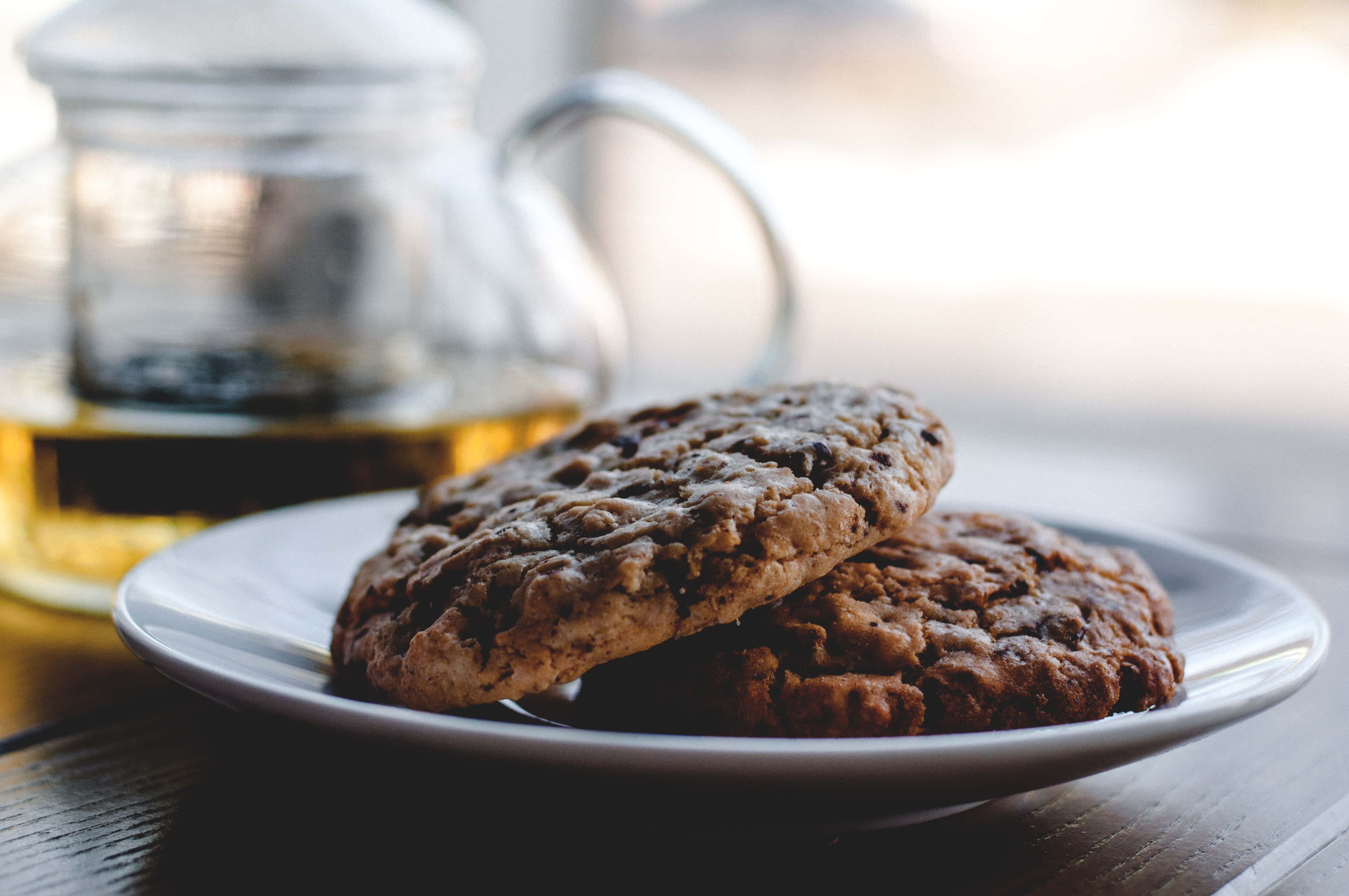 Cookie 桌布 壁紙 — 兩塊溫暖的巧克力餅乾放在白色盤子上，旁邊有一個裝滿金黃色茶的玻璃茶壺。