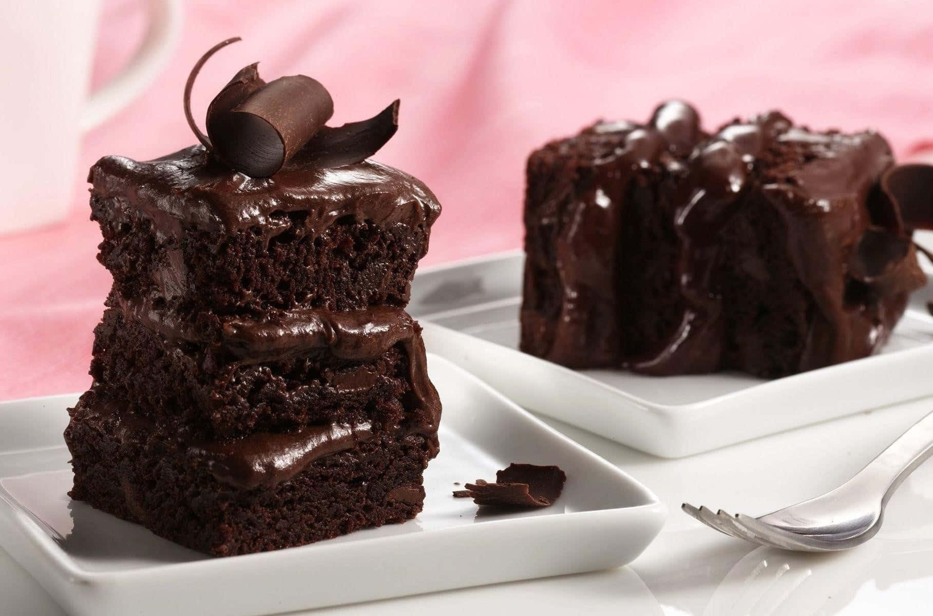 Chocolate Cake 桌布 壁紙 — 兩塊濃郁的巧克力蛋糕片，帶有光澤的甘納許和捲曲，放在白色盤子上，背景是柔和的粉紅色。
