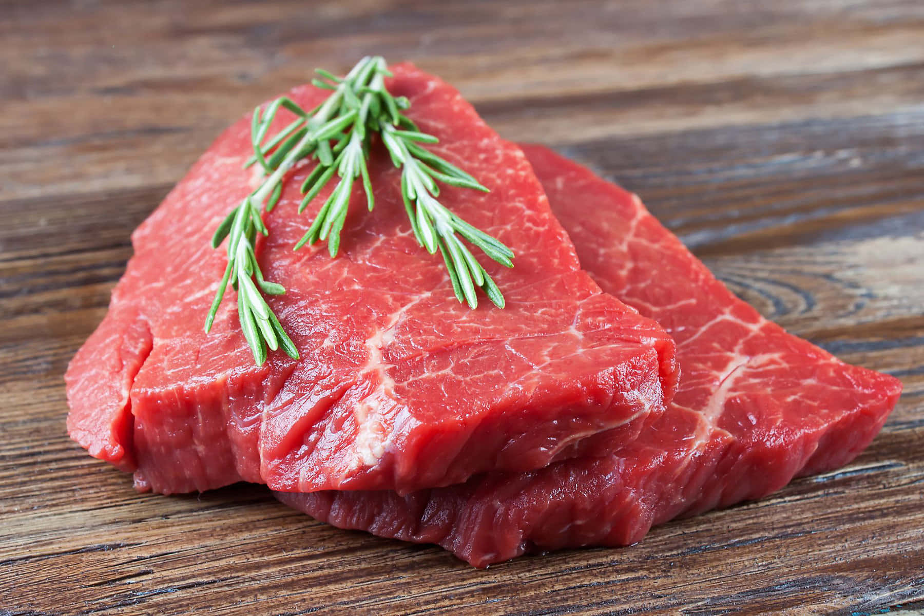 Red Meat 桌布 壁紙 — 兩塊生牛肉排搭配迷迭香枝條，放置在質樸的木質表面上。