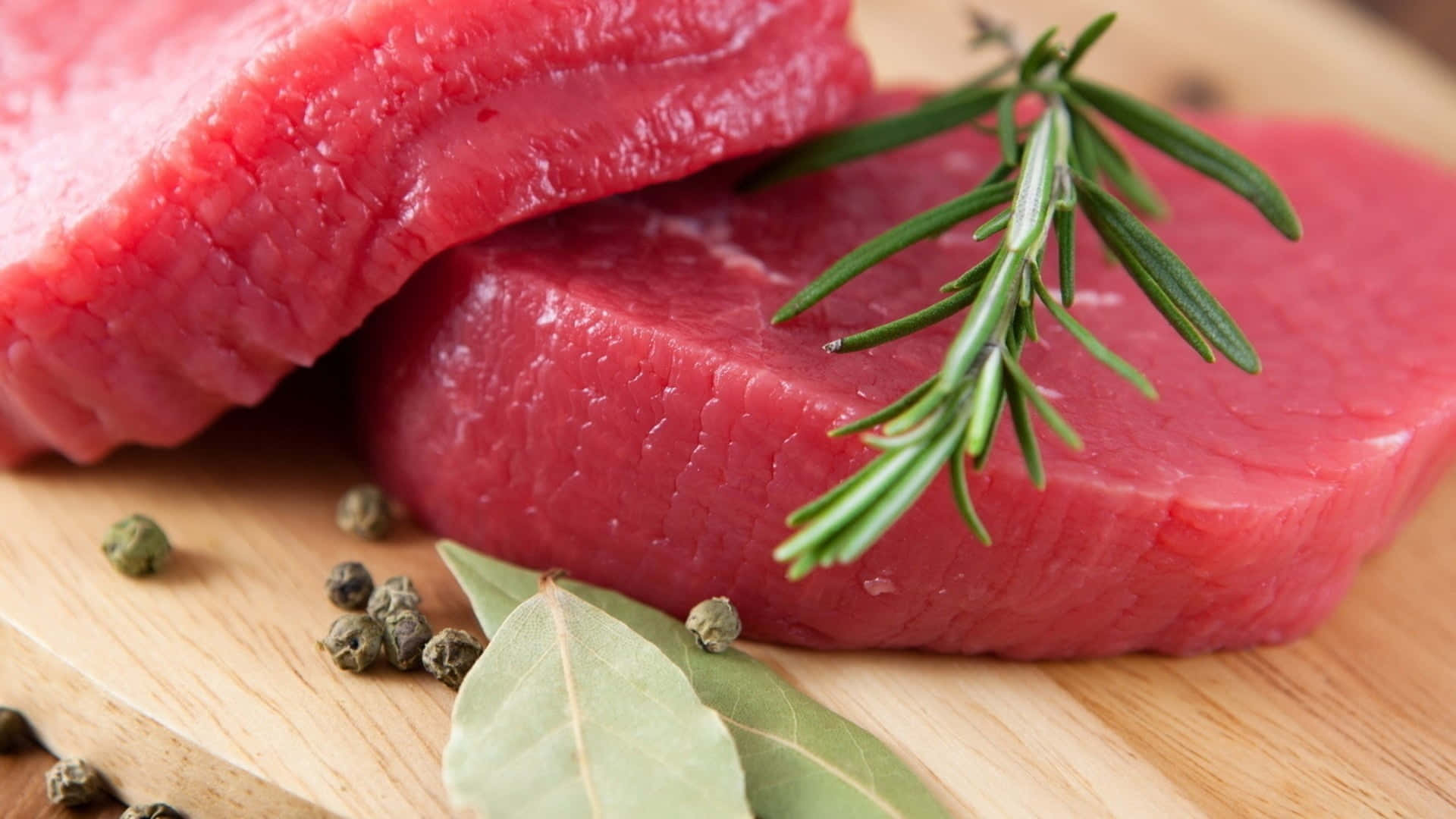 Red Meat 桌布 壁紙 — 兩塊生牛肉排放在木板上，以迷迭香、月桂葉和胡椒粒裝飾。