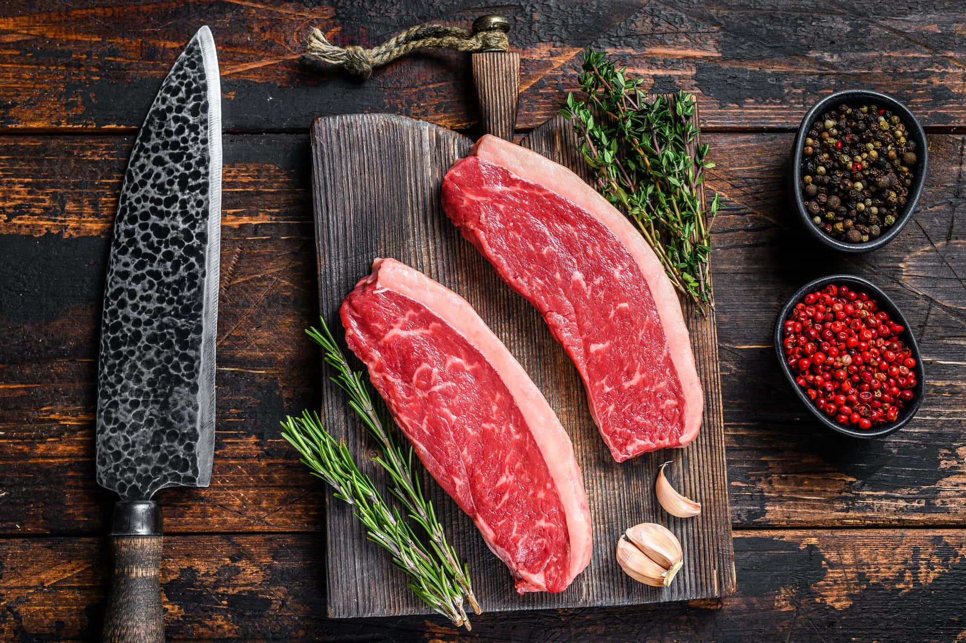 Red Meat 桌布 壁紙 — 兩塊生牛肉牛排放在木製砧板上，旁邊是新鮮香草、大蒜、胡椒粒和一把錘紋刀，置於質樸的深色木質表面上。