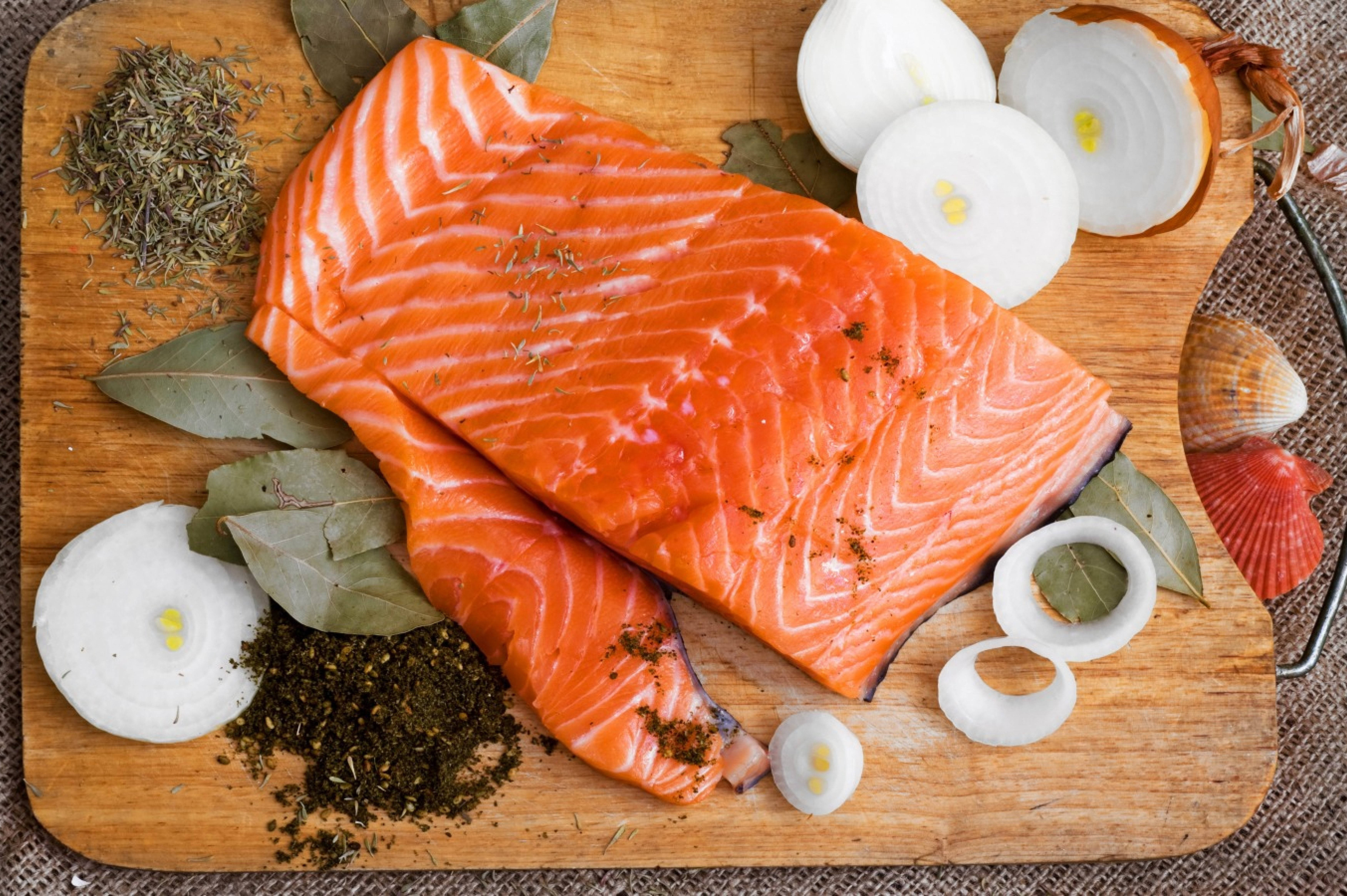 Delicious Food 桌布 壁紙 — 兩塊生鮭魚片，帶有橙黃色大理石紋的魚肉，放在木砧板上，周圍有洋蔥片、月桂葉和乾燥香草。