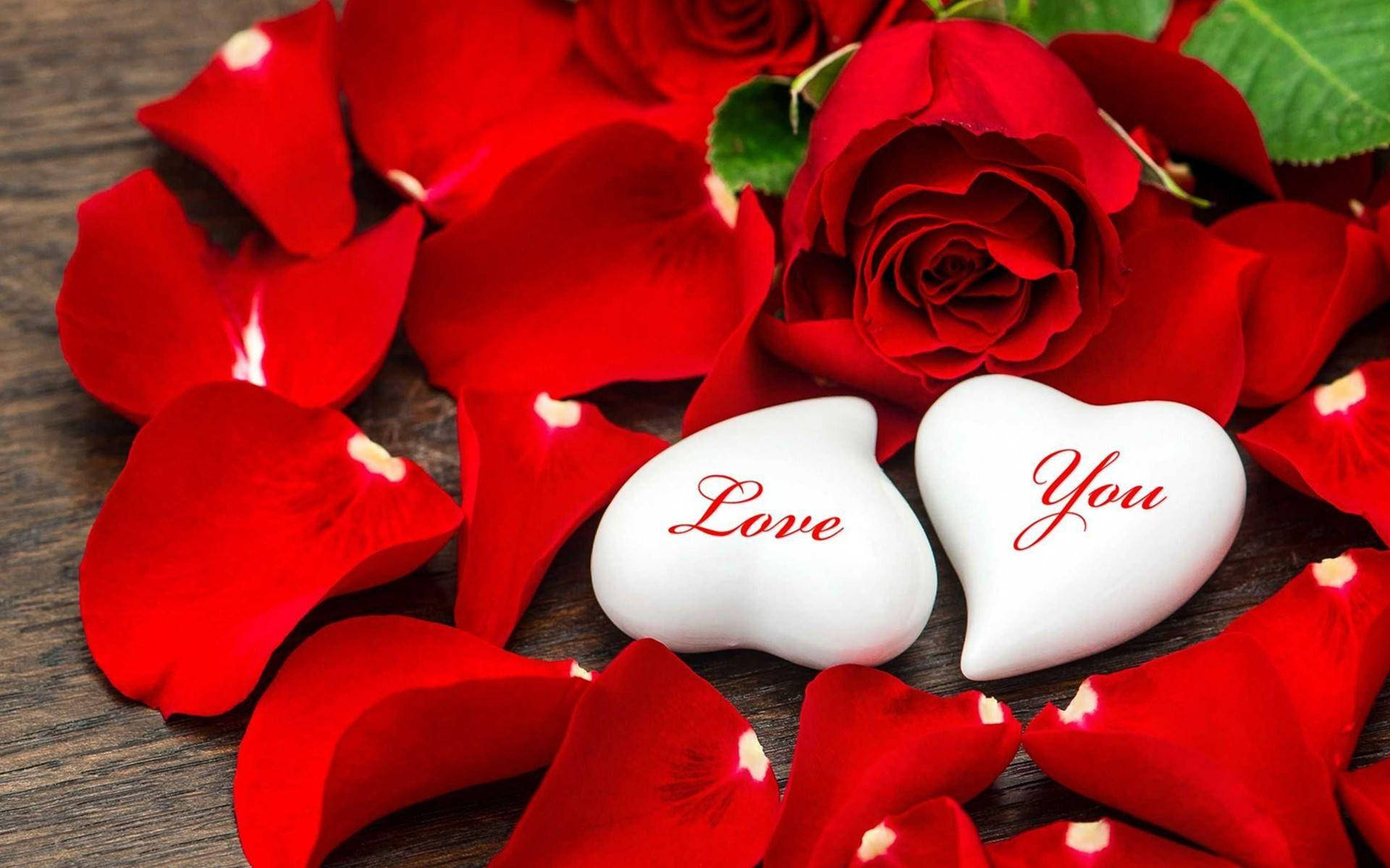 Red Rose Aesthetic 桌布 壁紙 — 兩塊白色心形石頭刻有「Love You」，鑲嵌在鮮紅玫瑰花瓣中，置於質樸的木質表面上。