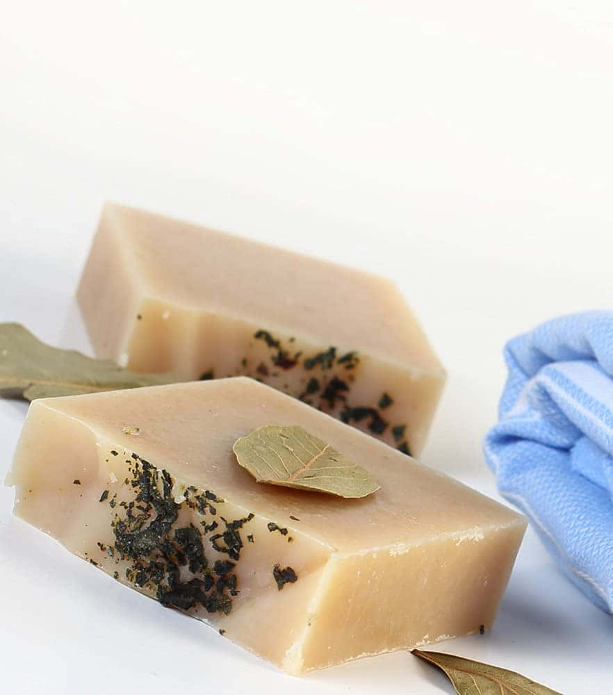 bath 桌布 壁紙 — 兩塊米色手工皂，內嵌深色草本和一片月桂葉，放在白色表面上，旁邊有一條折疊的藍色毛巾。