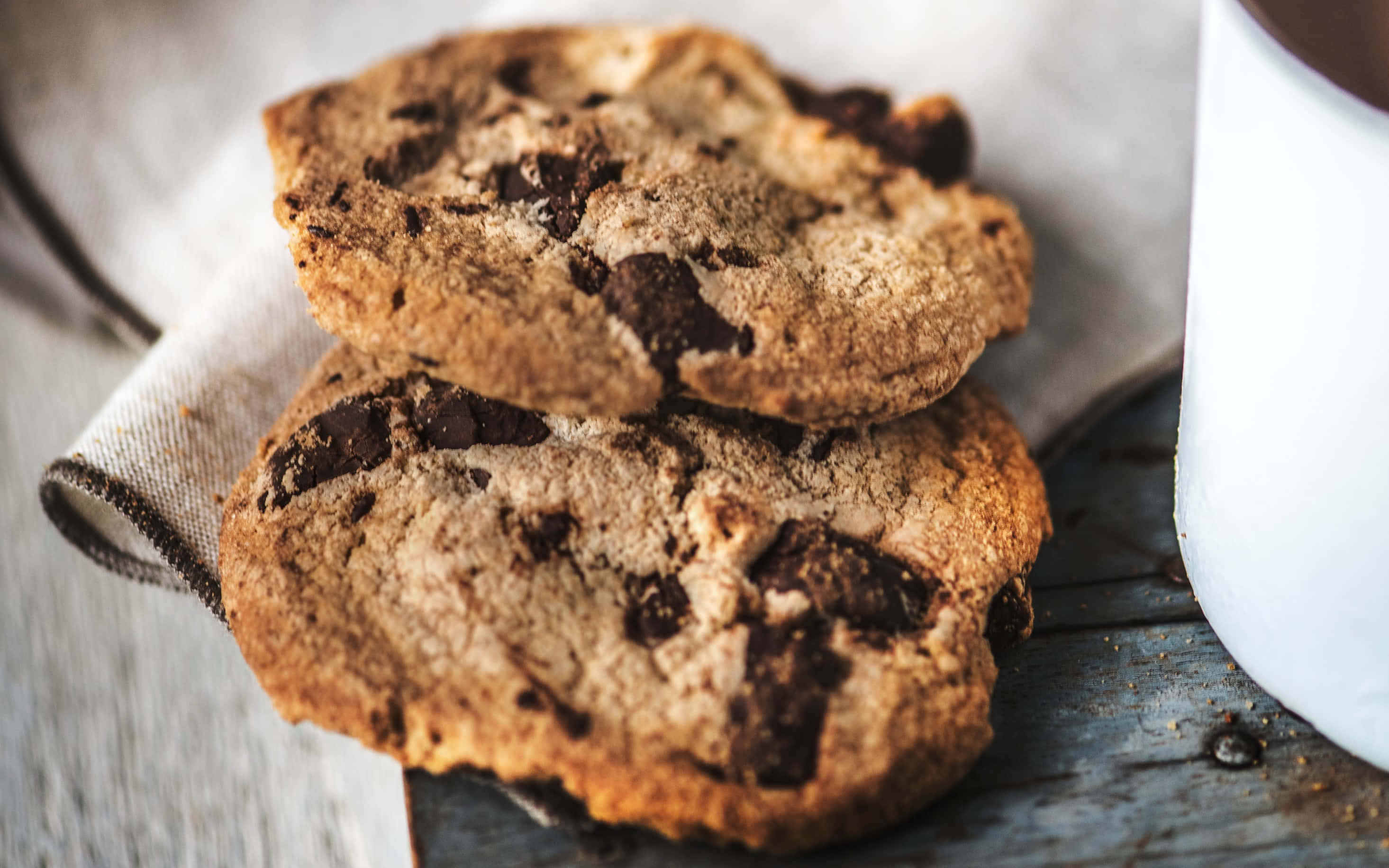 Cookie 桌布 壁紙 — 兩塊金黃色的巧克力餅乾放在一塊布上，旁邊有一個白色馬克杯，置於鄉村風格的木質表面上。