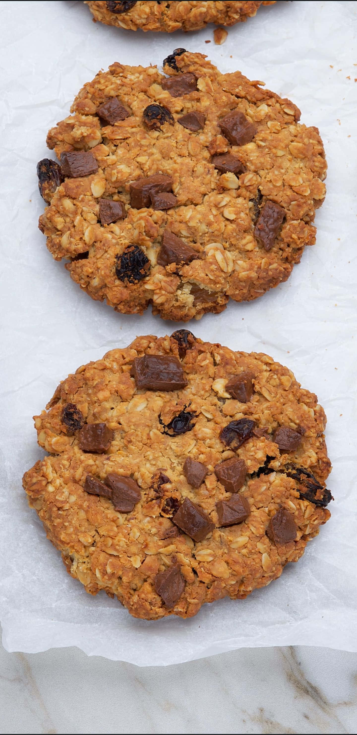 Oatmeal 桌布 壁紙 — 兩塊金黃色的燕麥餅乾，上面鑲有巧克力塊和葡萄乾，放在烘焙紙上。