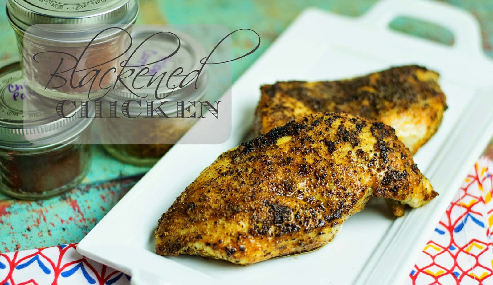 Blackened Chicken 桌布 壁紙 — 兩塊金黃色的黑雞胸肉放在白色砧板上，旁邊是放在彩色圖案布上的香料罐。