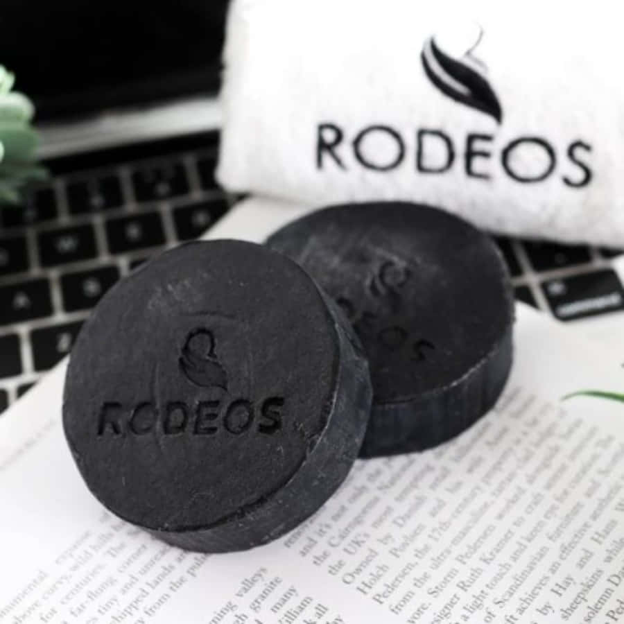 Black Soap 桌布 壁紙 — 兩塊霧面黑色圓形RODEOS肥皂放在一本打開的書上，旁邊有品牌毛巾和筆記型電腦鍵盤。