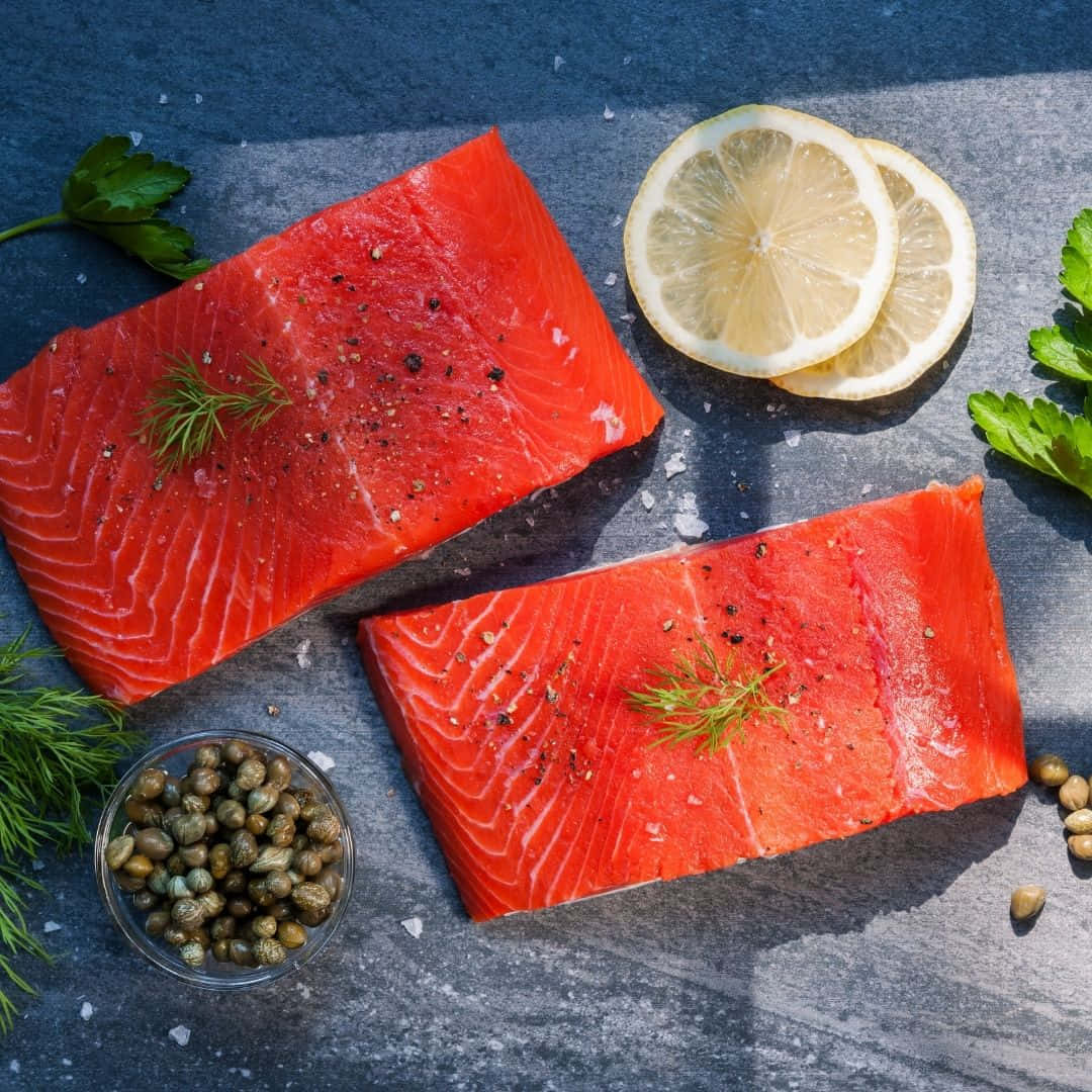 Salmon 桌布 壁紙 — 兩塊鮮紅的鮭魚菲力，以鹽、胡椒和蒔蘿調味，放在深色石板表面上，旁邊有檸檬片、酸豆和新鮮香草。